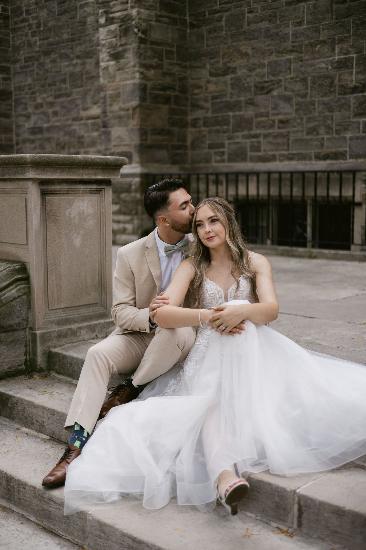 Groom tenderly kisses bride's temple as they sit on stone steps outside a historic stone building.