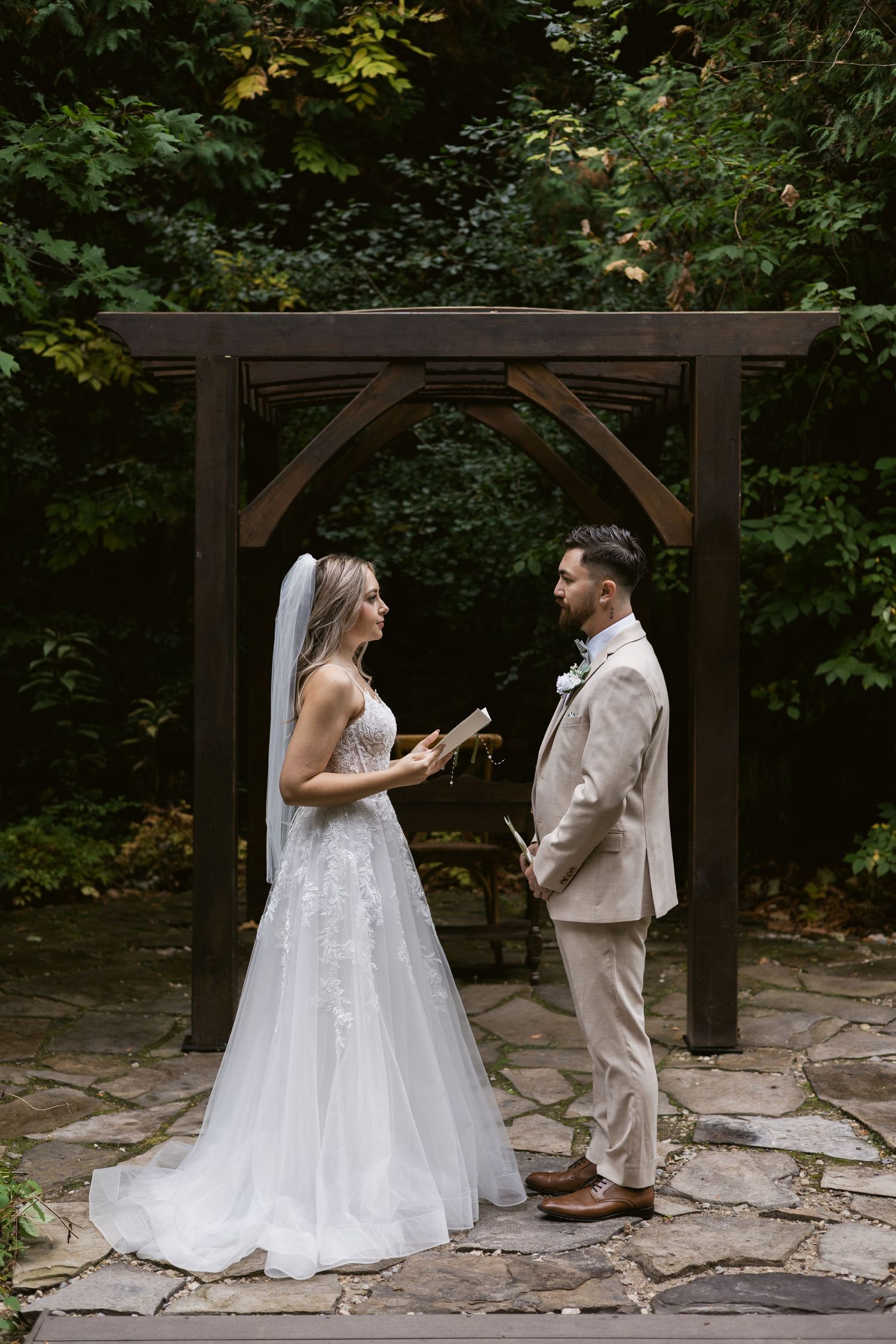 Bride and groom facing each other during intimate outdoor vow exchange under rustic wooden arch.