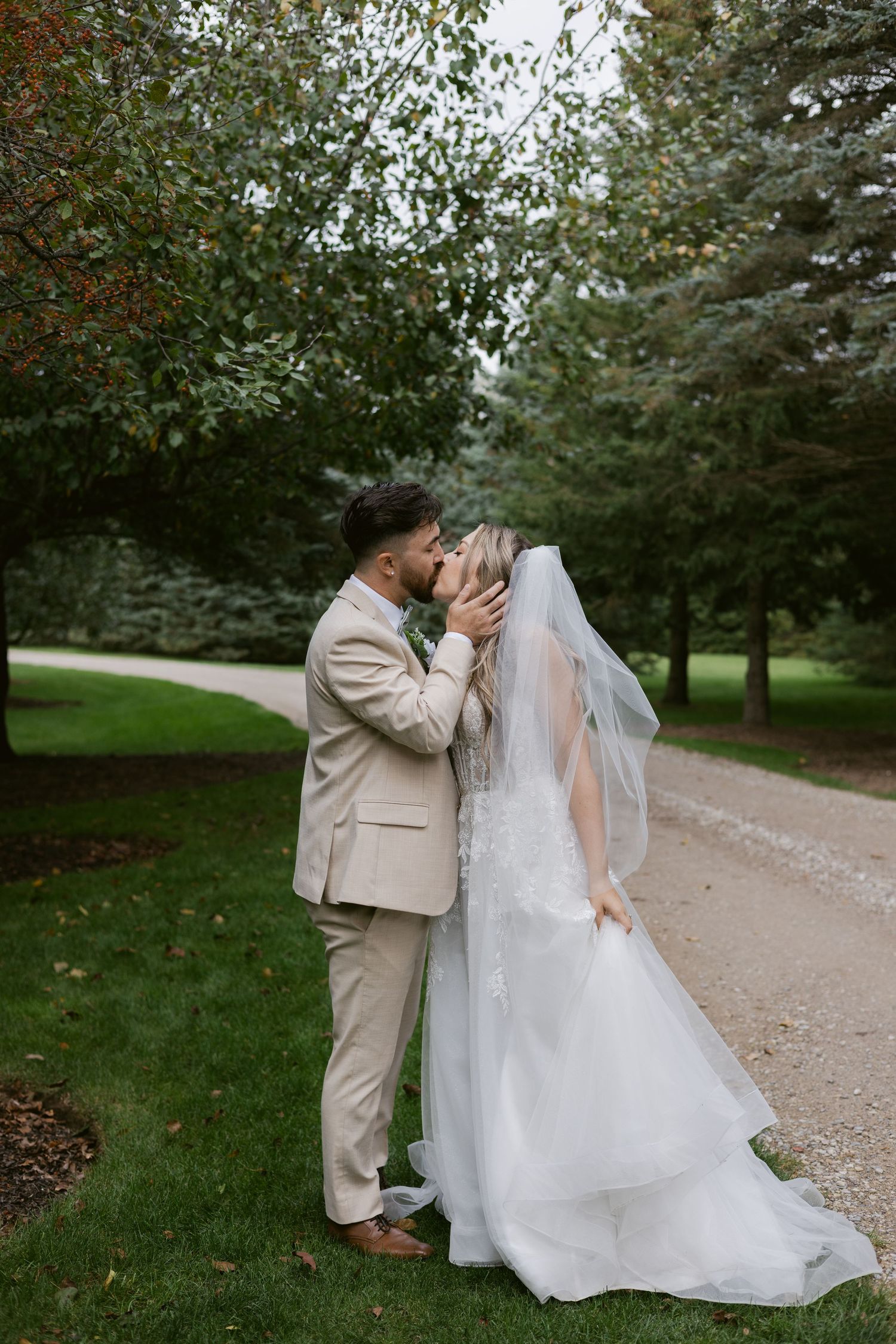 Bride and groom share a romantic kiss outdoors on gravel path, bride in white lace gown with flowing veil.