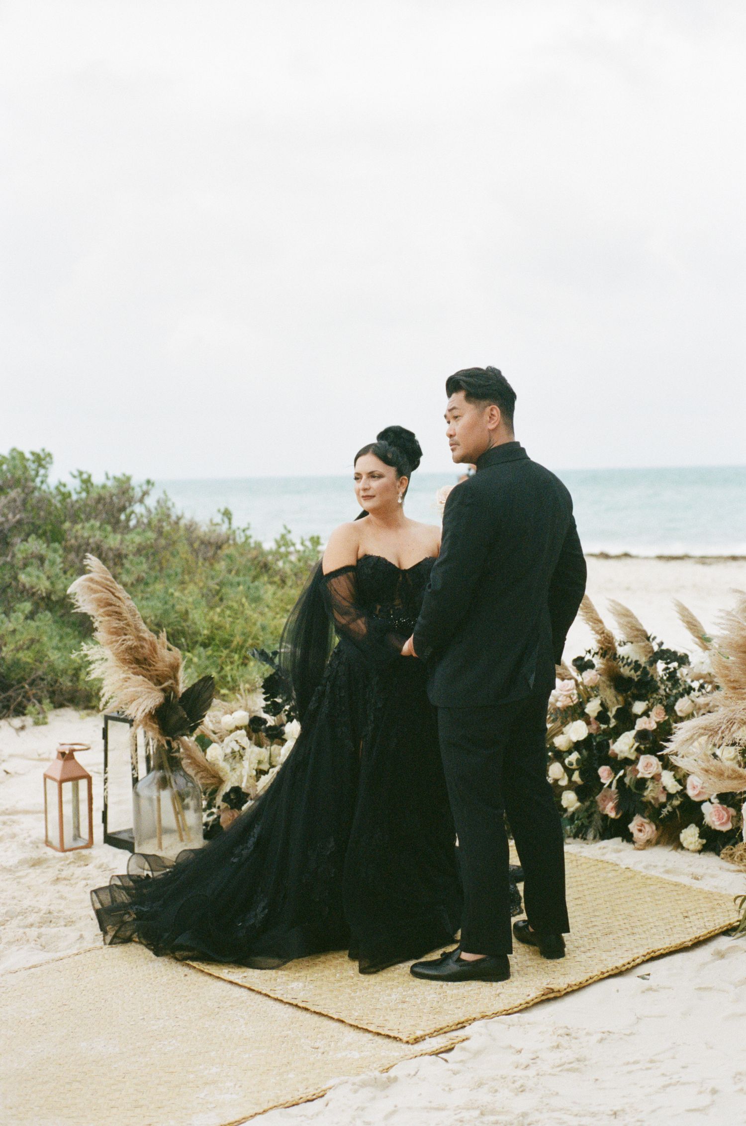 Elegant couple in black wedding attire at a beach ceremony altar surrounded by pampas grass and white roses.