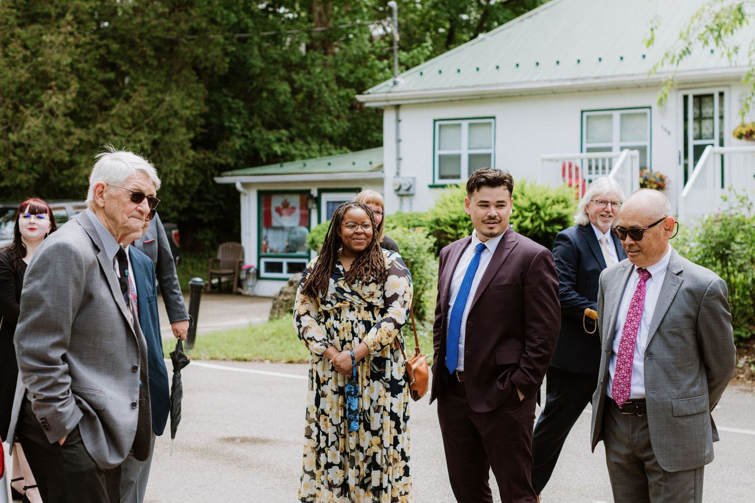Wedding guests in formal attire walk together outside a white building surrounded by trees.