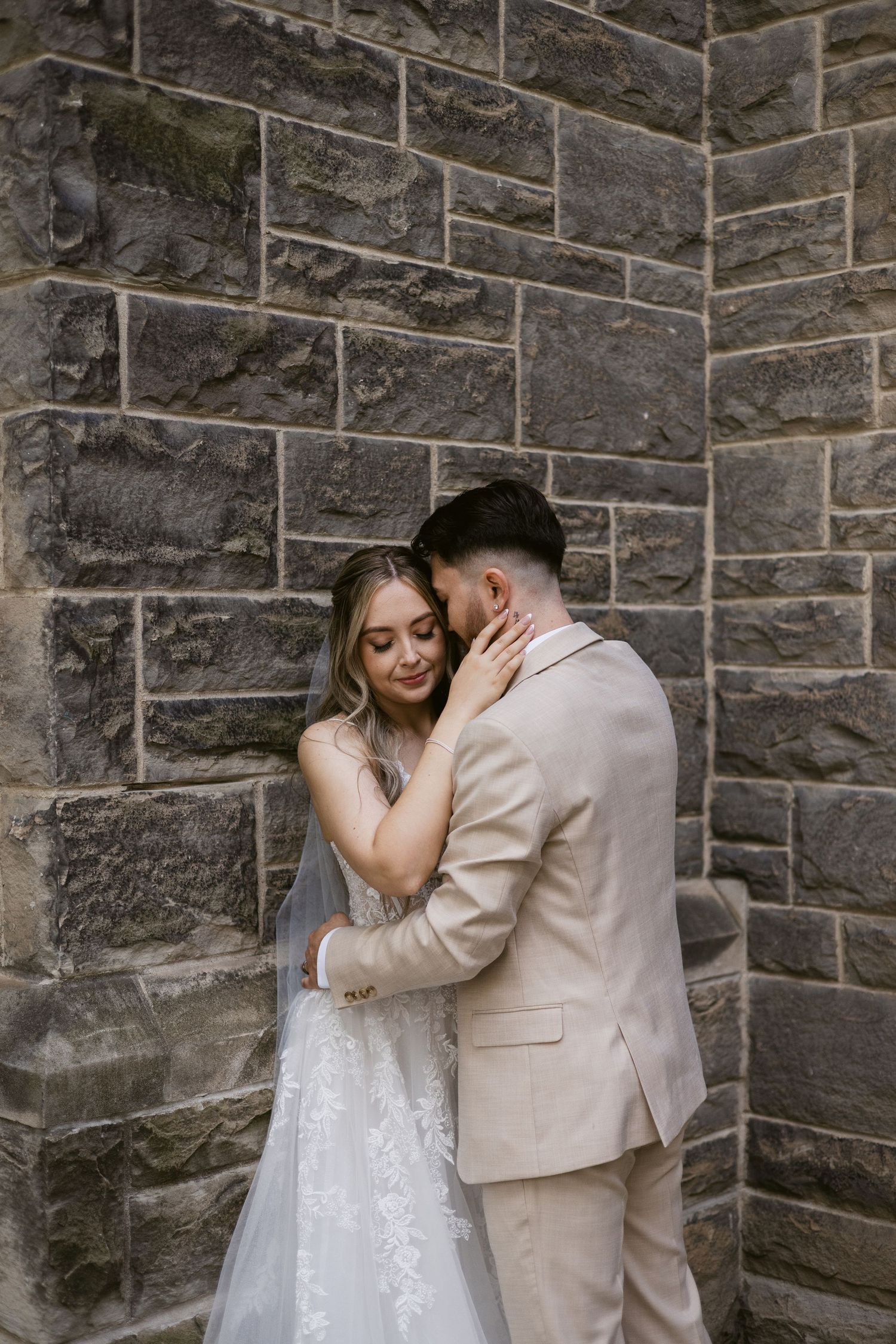 Bride gently holds grooms face as they embrace against a textured stone wall in romantic wedding portrait.