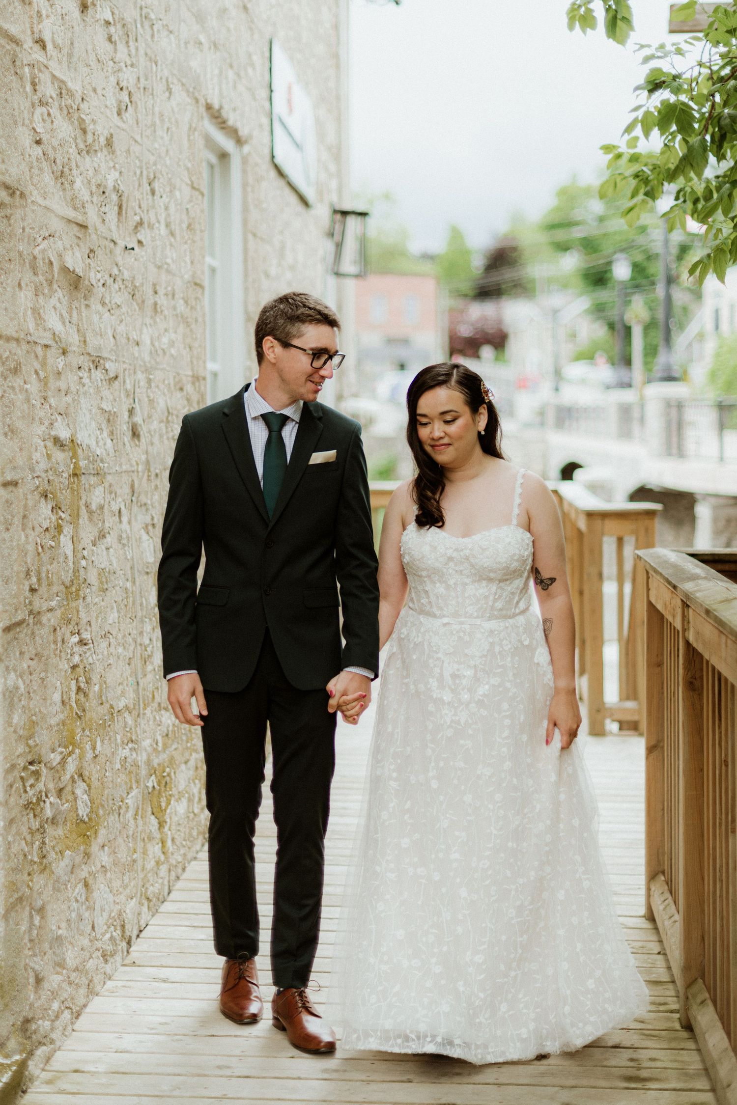 Bride and groom holding hands walking along wooden boardwalk beside stone building on wedding day.