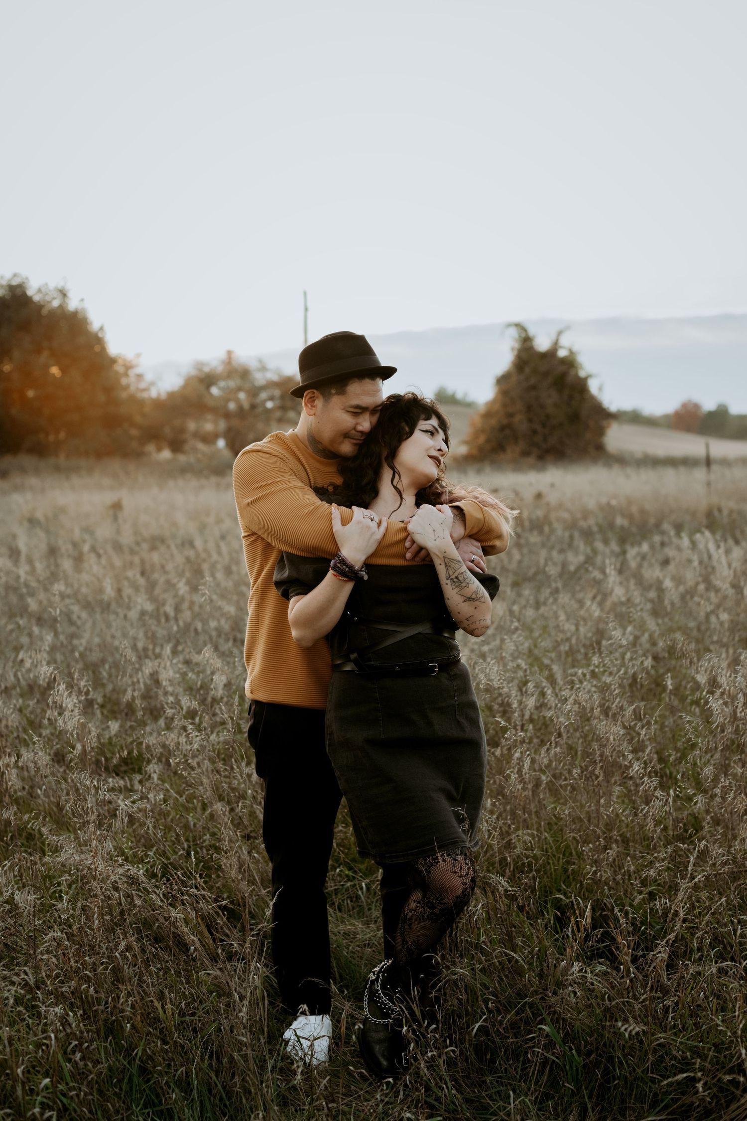 Couple sharing a tender embrace in autumn field at sunset, man in black hat and mustard sweater with tattooed woman.