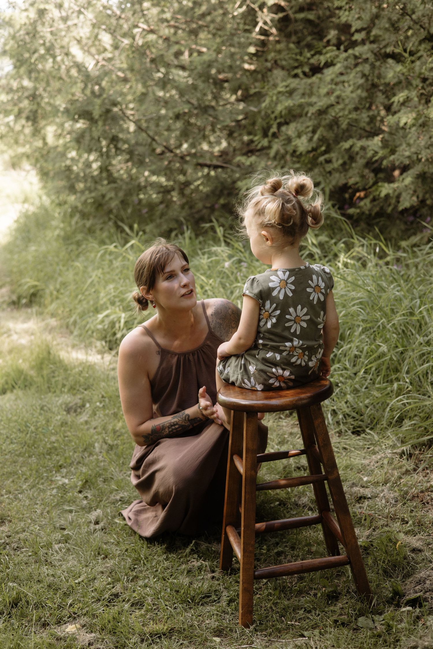 Tattooed mother in brown dress kneels on grass speaking to toddler daughter sitting on wooden stool outdoors.