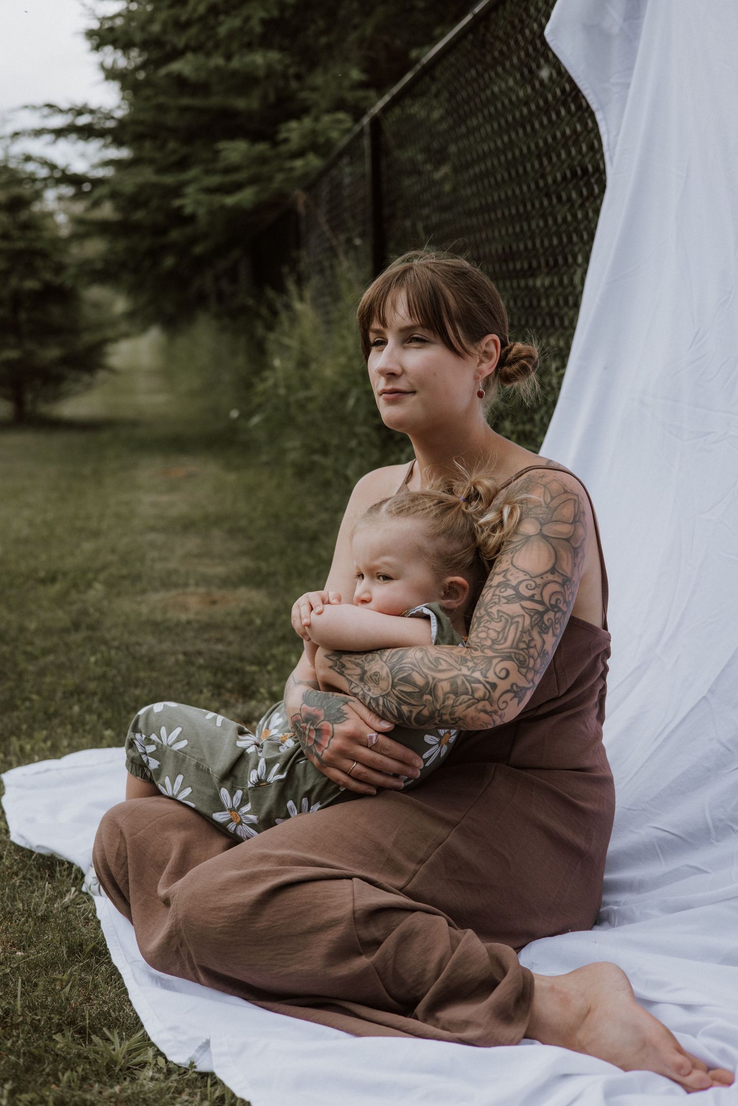 A tattooed mother gently embraces her sleepy toddler while sitting on a white blanket outdoors.