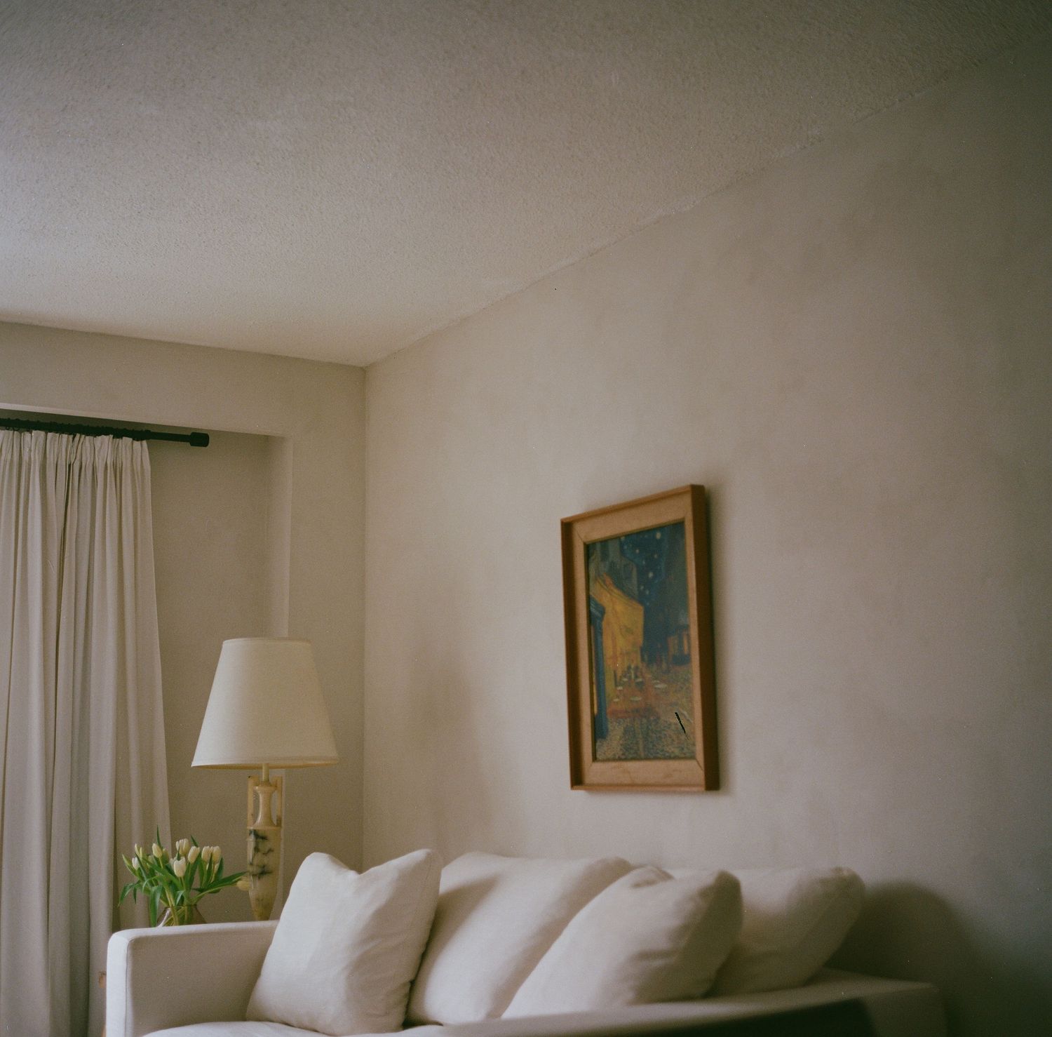 Minimalist living room with white sofa, cream curtains, table lamp, and Van Gogh Cafe Terrace framed print.