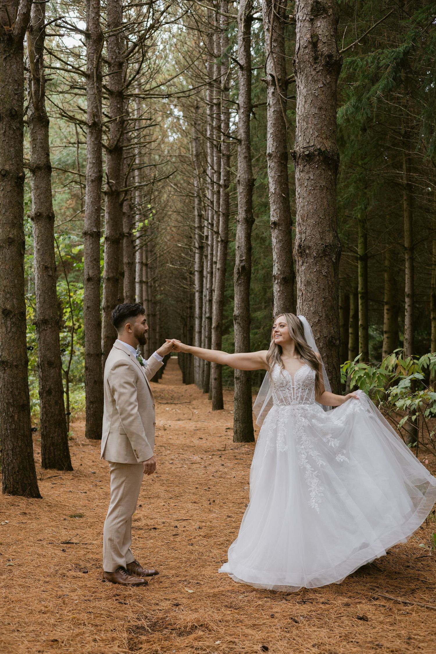 Bride and groom dancing in a pine forest, bride in lace ballgown, groom in tan suit, holding hands.