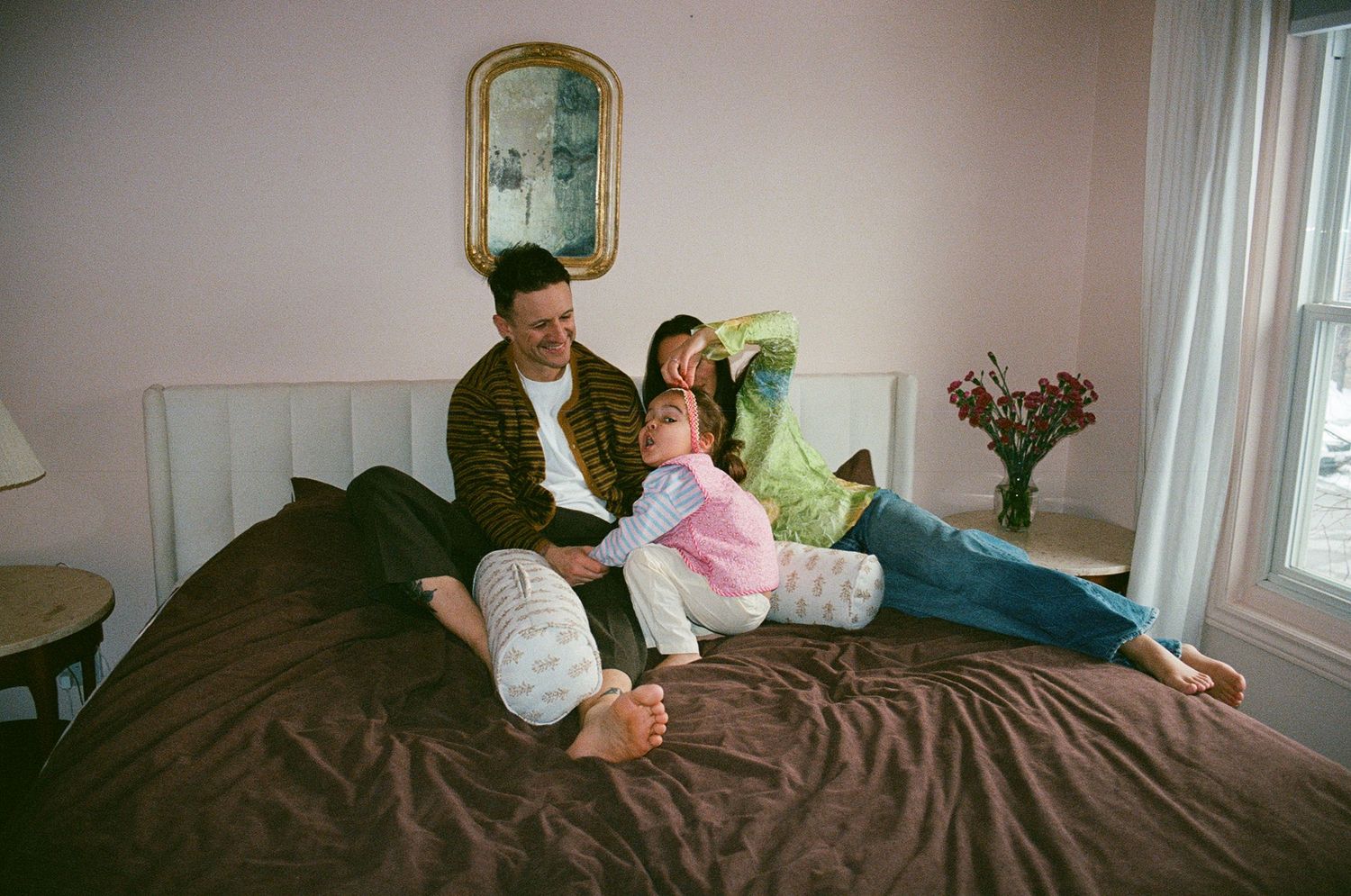 Couple relaxes playfully on brown bedding, woman in green top and jeans, man in striped cardigan, pink walls.