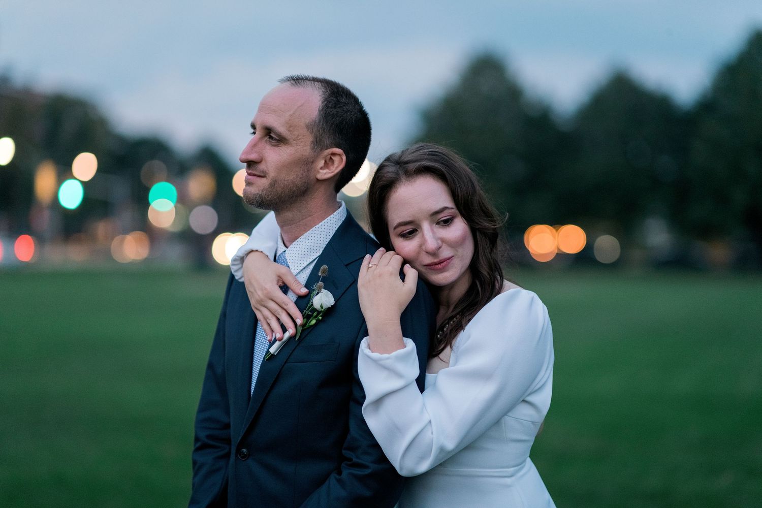 Bride resting her head on groom's shoulder at dusk with bokeh city lights in background.
