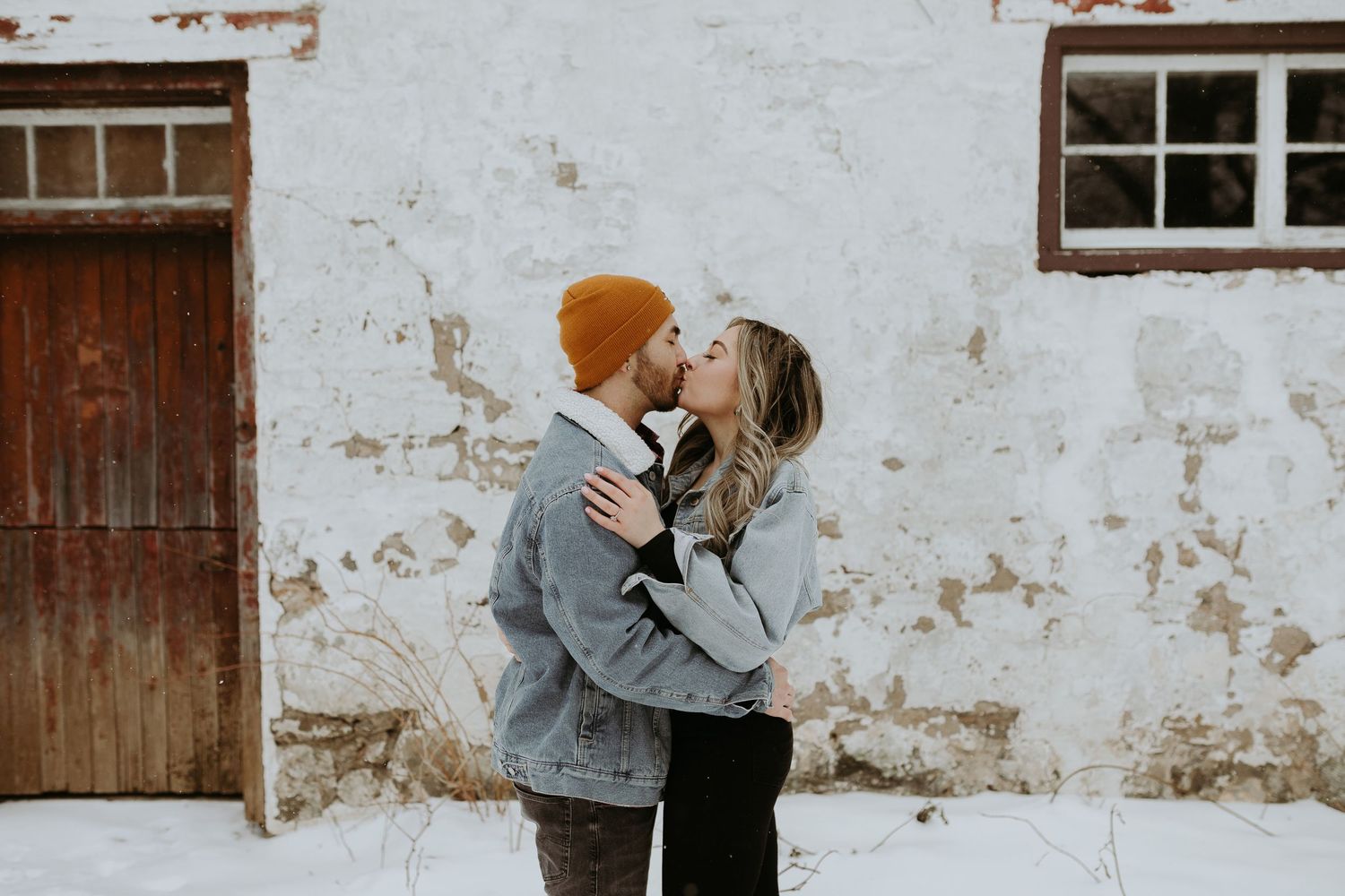 Romantic couple kissing against rustic white weathered wall in snowy winter setting.