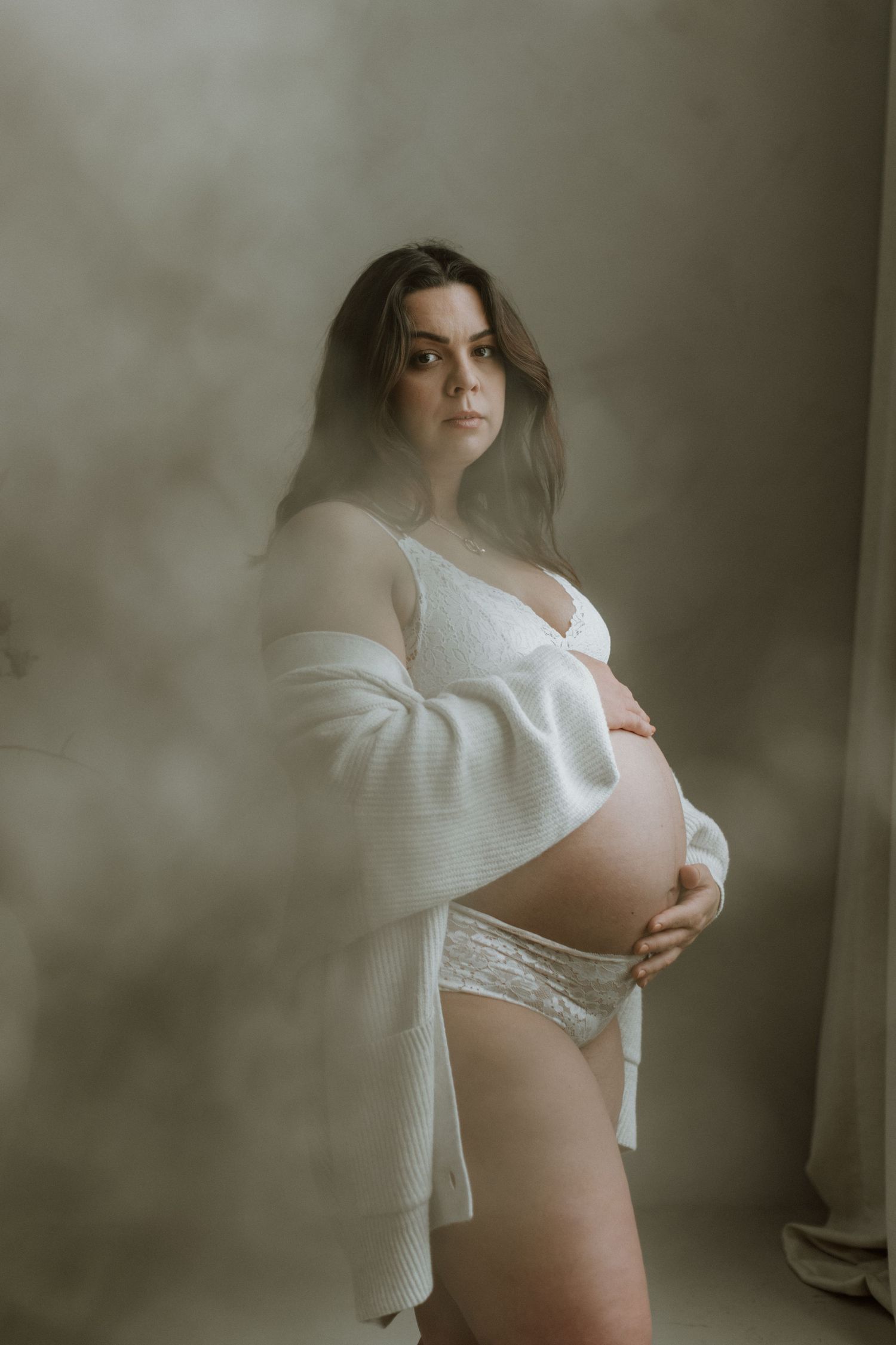 Pregnant woman in white lace lingerie and open cardigan gently holding her bare baby bump.