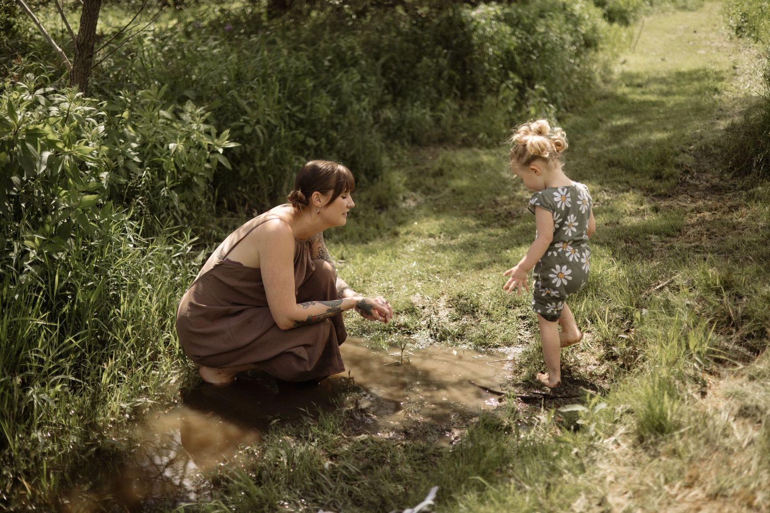 Mother in brown dress kneels by stream with toddler girl in daisy romper in lush green nature.