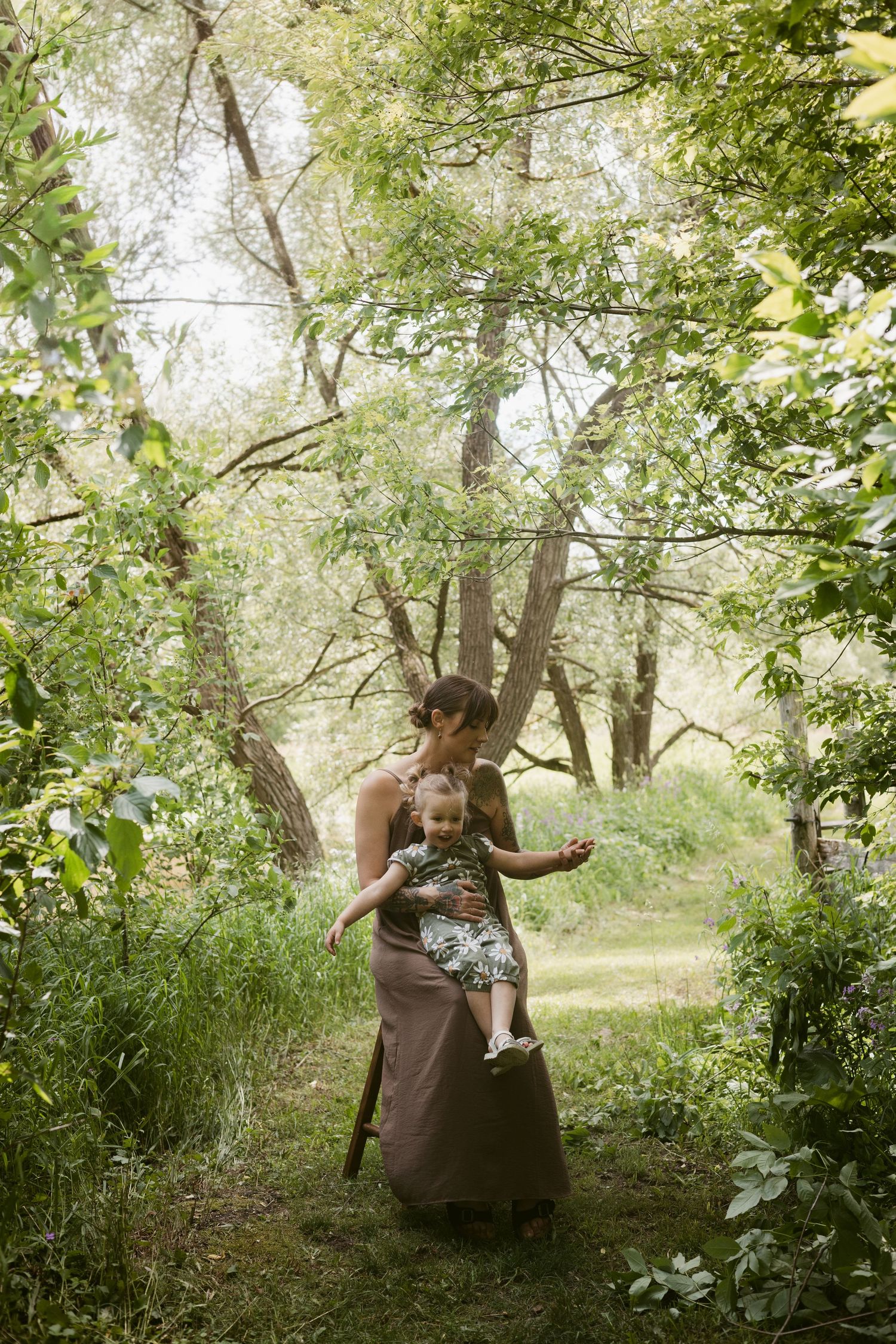 A tattooed mother carries her young daughter through a lush green wooded area outdoors.
