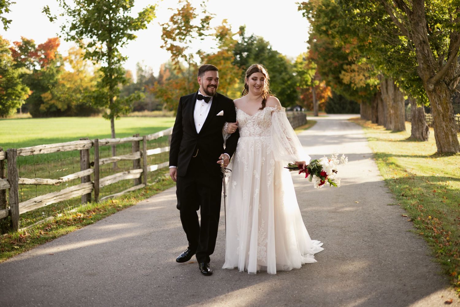 Bride and groom walking hand in hand along a tree-lined path in autumn, bride in white gown.