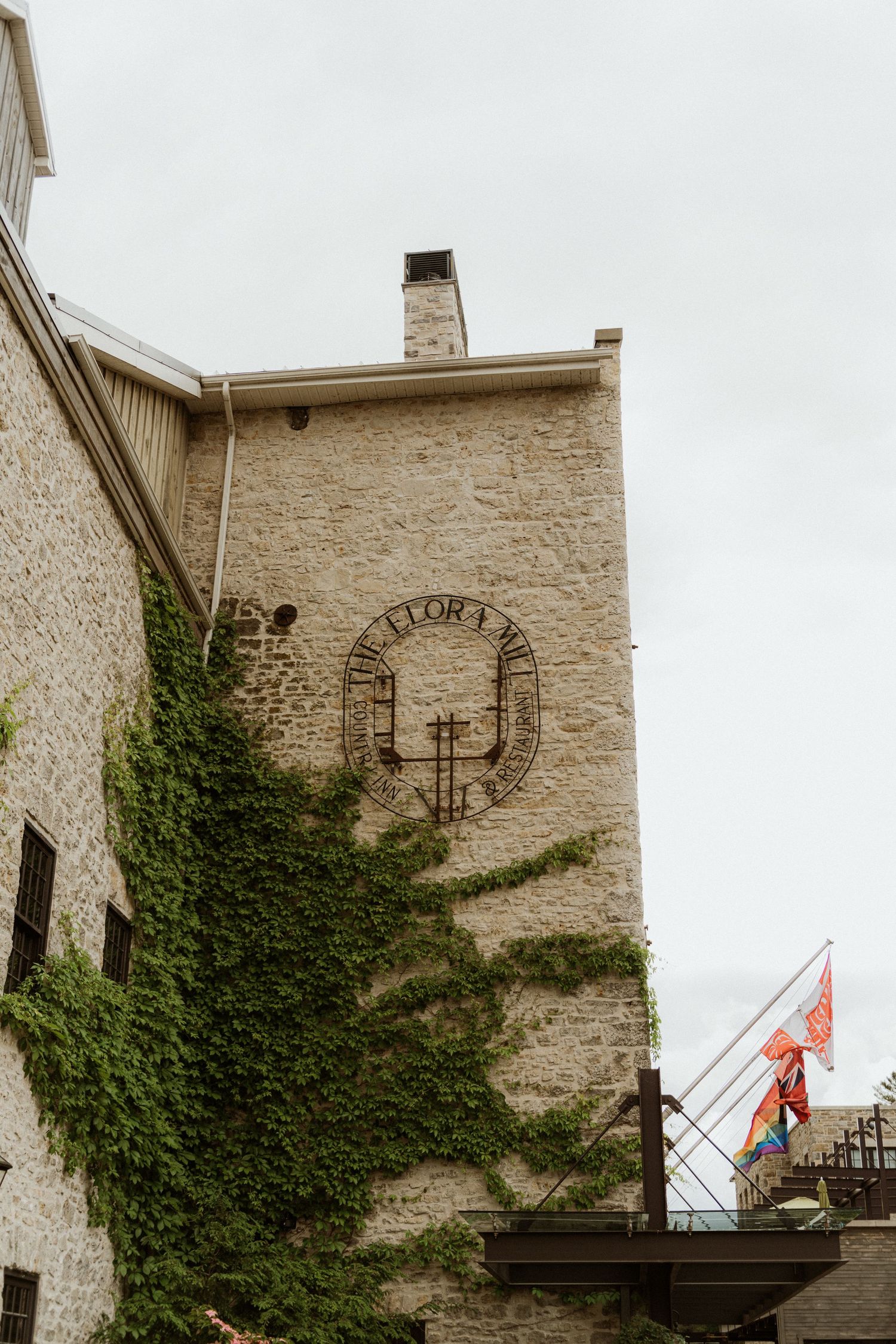 Ivy-covered limestone wall of Elora Mill with circular logo and pride flags on balcony.