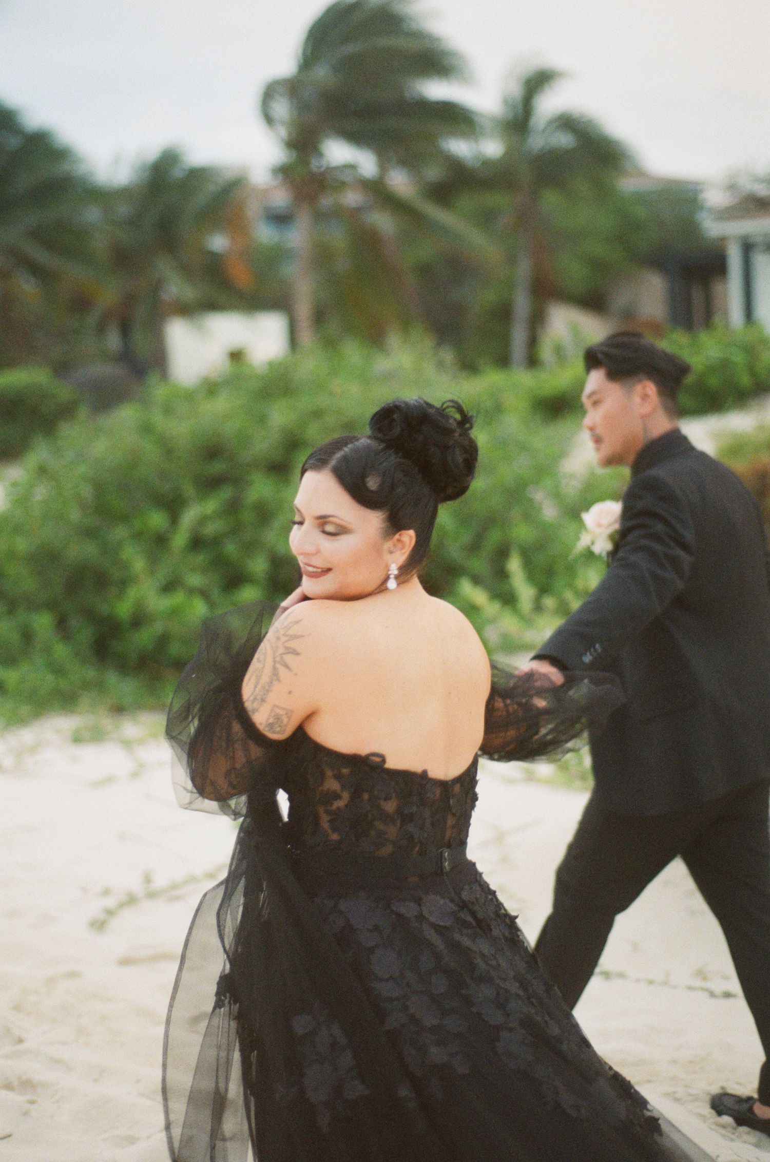 Bride in black lace strapless gown with tulle sleeves and tattoo dances on beach.