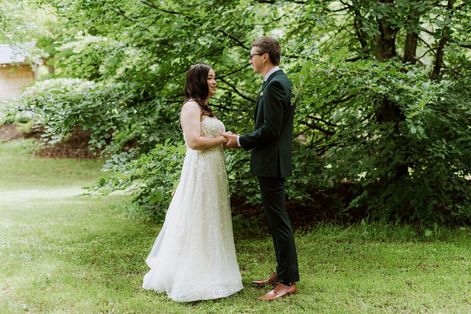 Bride and groom hold hands facing each other during an outdoor first look in a lush green garden setting.