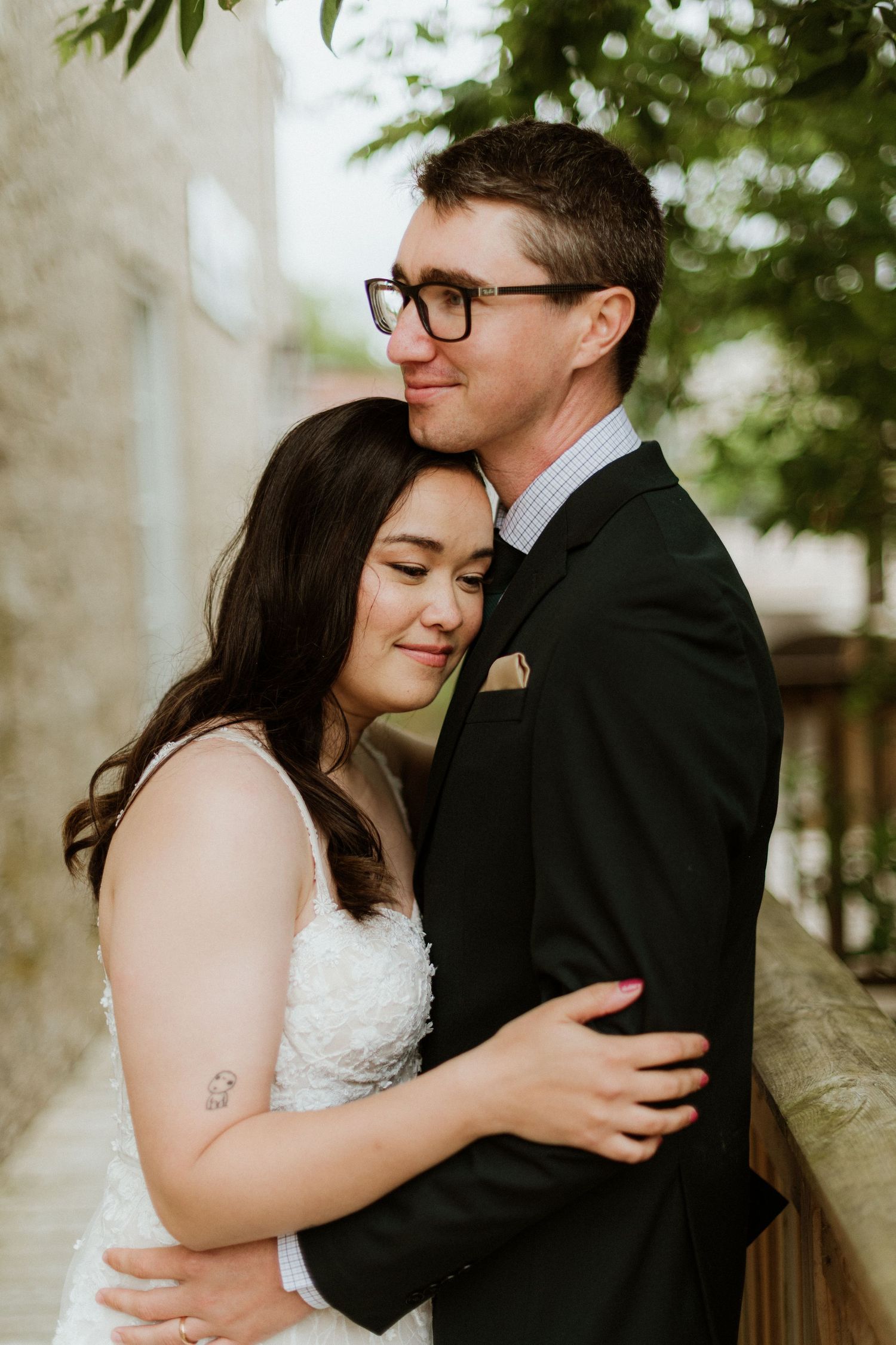 Bride in white lace dress embracing groom in black suit beneath green trees outdoors.
