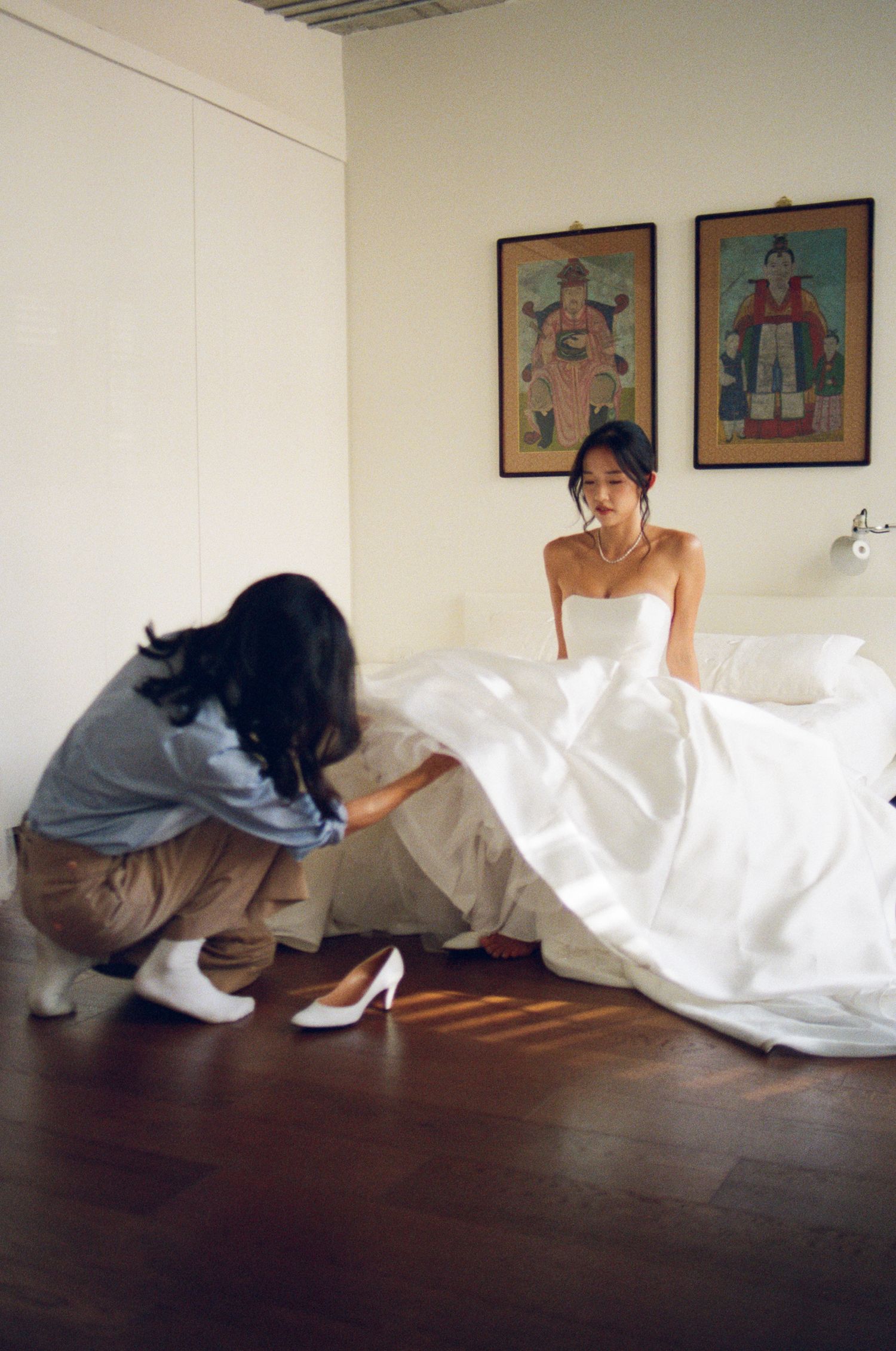 A bride in a strapless white gown sits on a bed while an attendant arranges her dress on the floor.