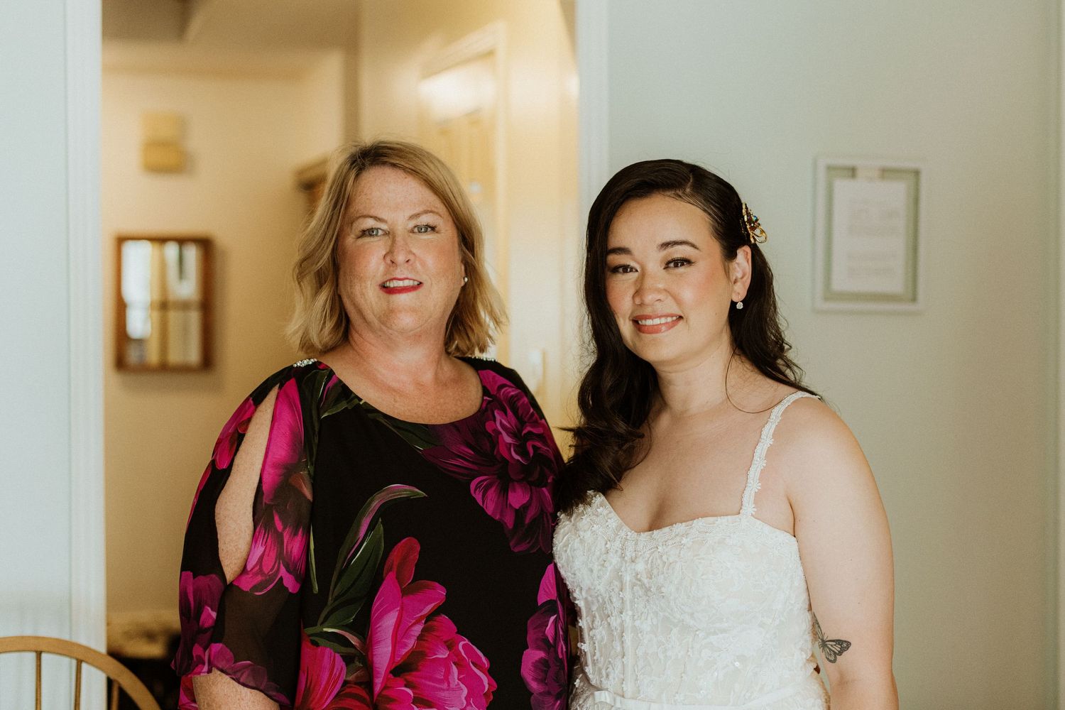 Smiling bride in white lace gown poses with older woman in floral dress on wedding morning.