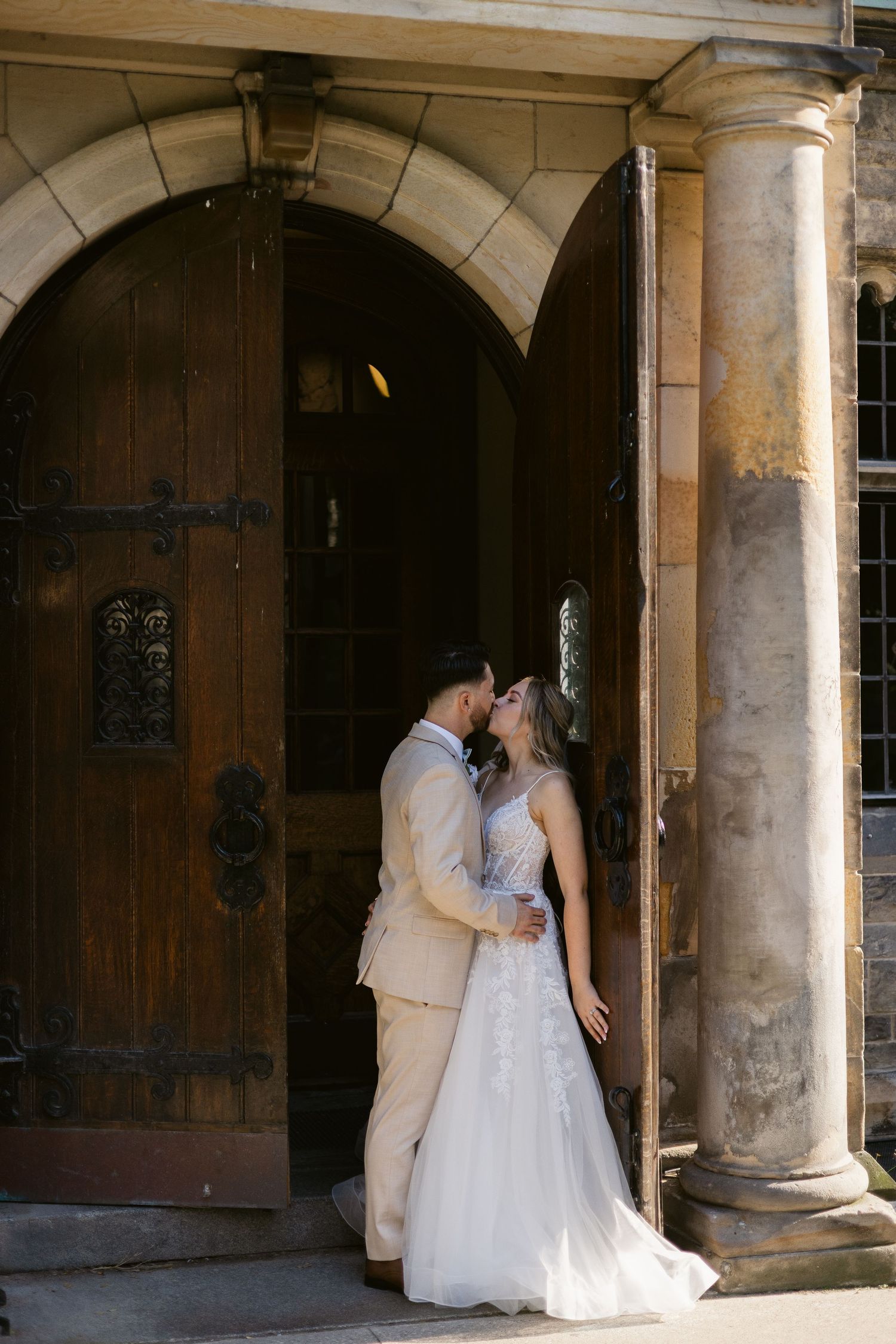 Bride and groom share a kiss framed by ornate wooden doors and stone columns of historic venue.