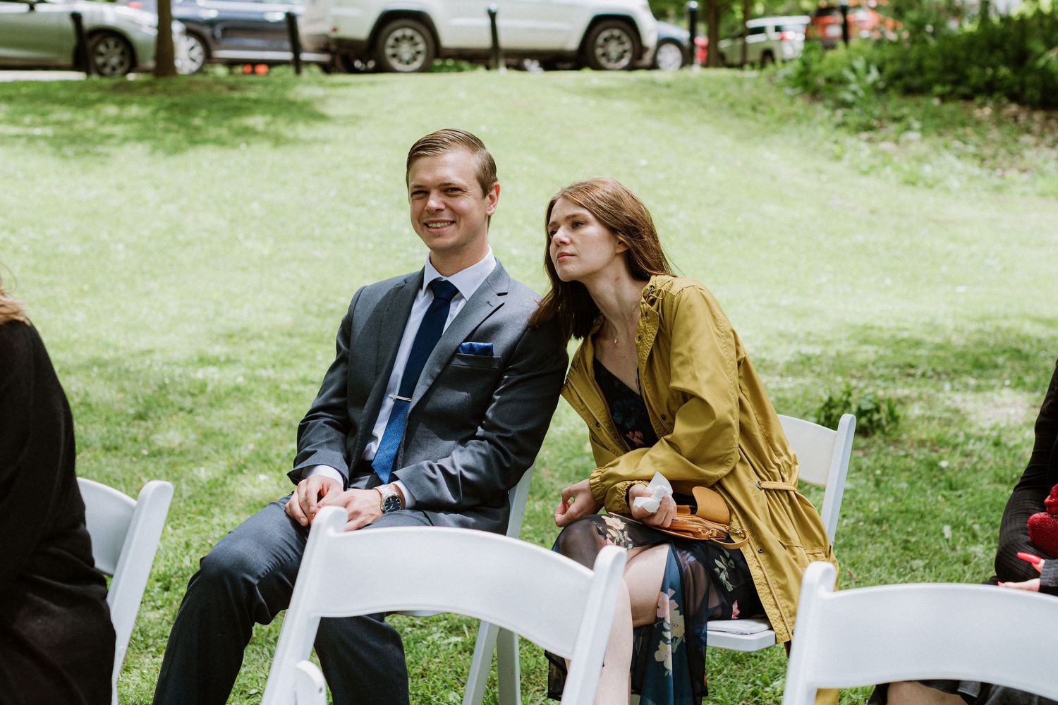 Wedding guests seated on white chairs outdoors, woman resting head on mans shoulder during ceremony.