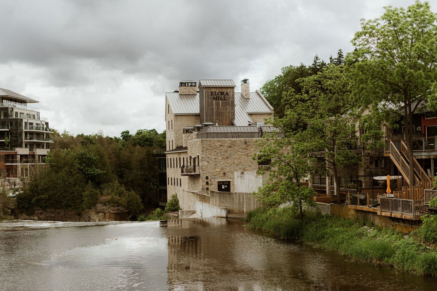 Historic stone mill building reflected in calm river waters surrounded by lush green trees.