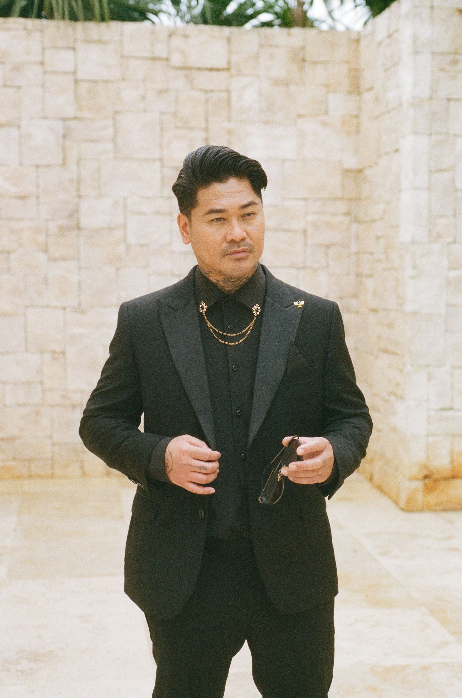 Groom in black tuxedo with gold chain necklace holds sunglasses against stone wall backdrop.
