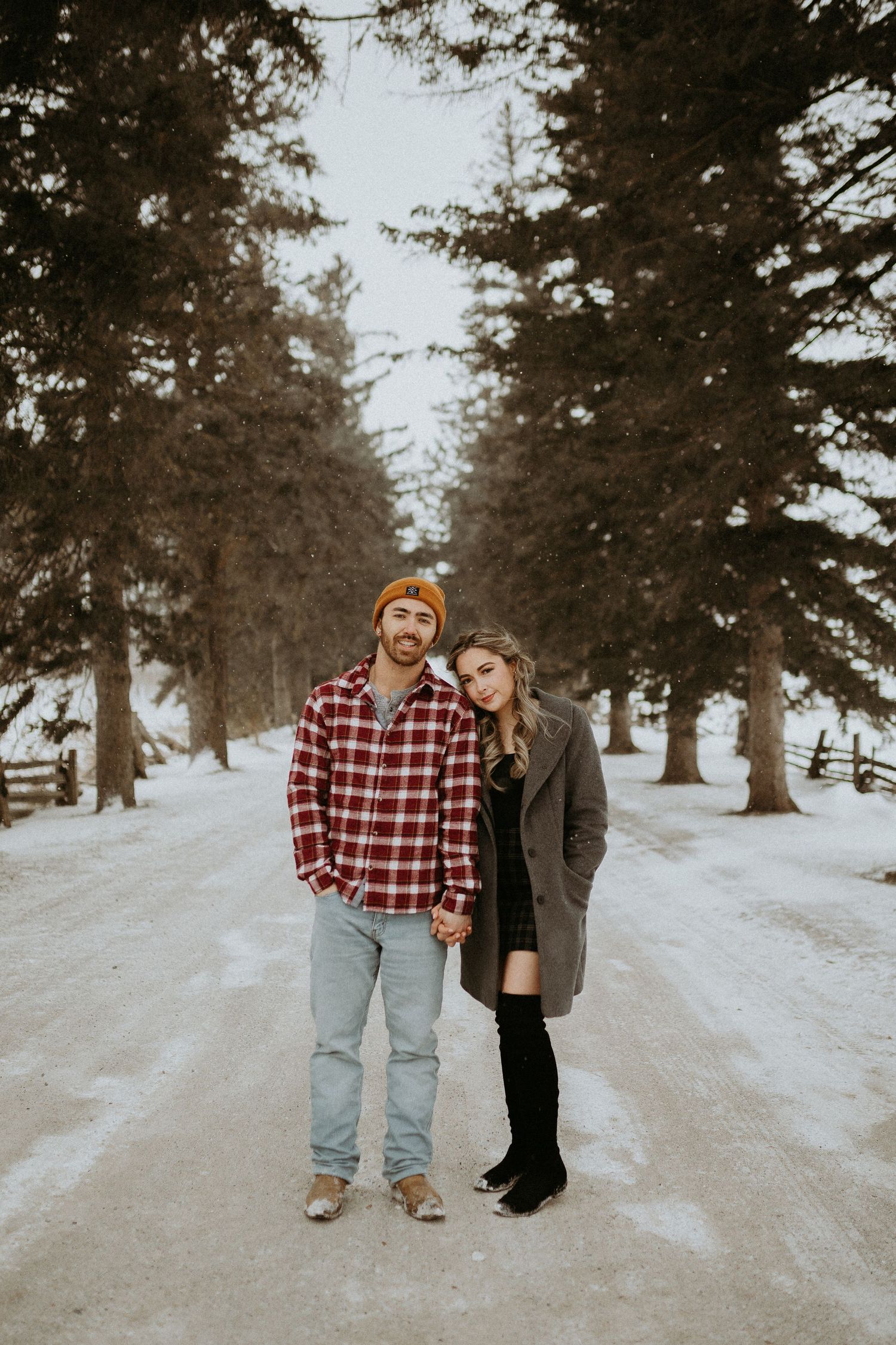 Couple posing together on snowy road surrounded by tall pine trees in winter.