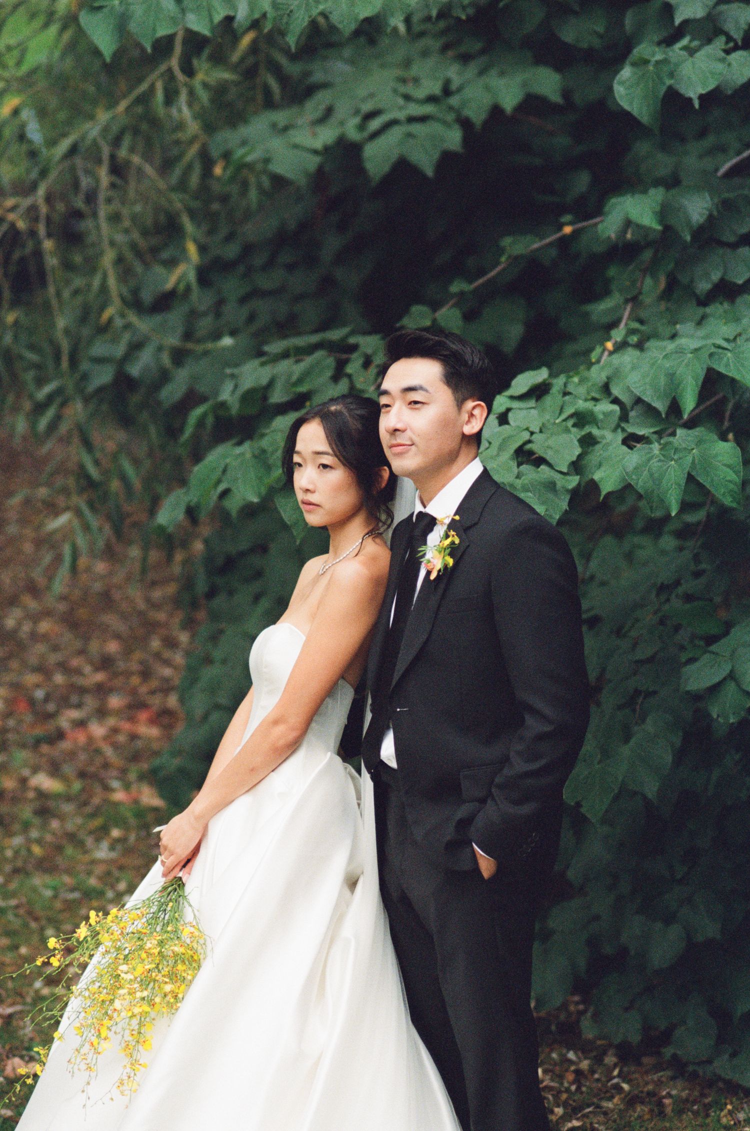 Asian bride and groom posing back-to-back before lush green foliage, bride holding yellow flowers.