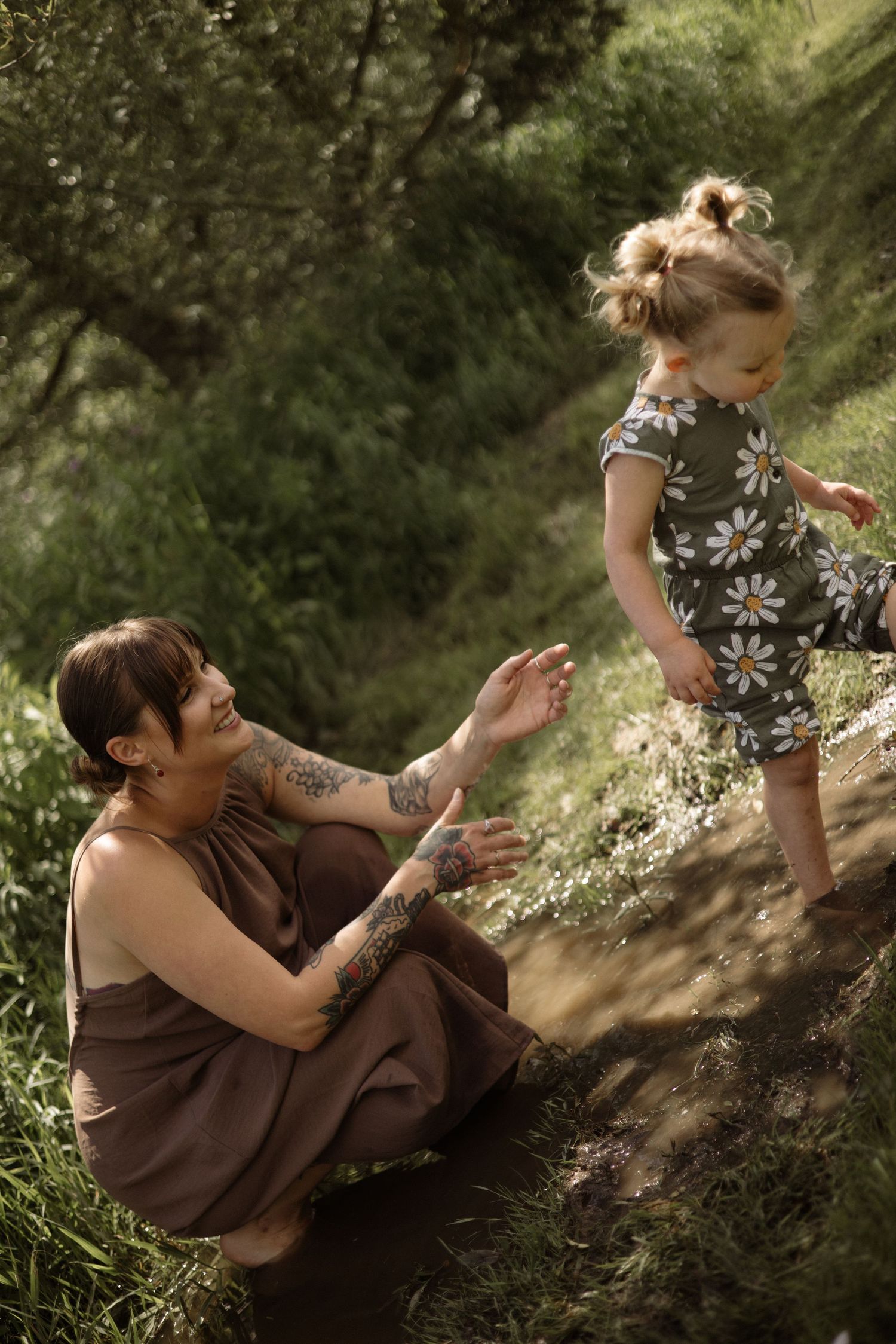 Tattooed mother in brown dress reaches for toddler girl in daisy romper on grassy hillside.