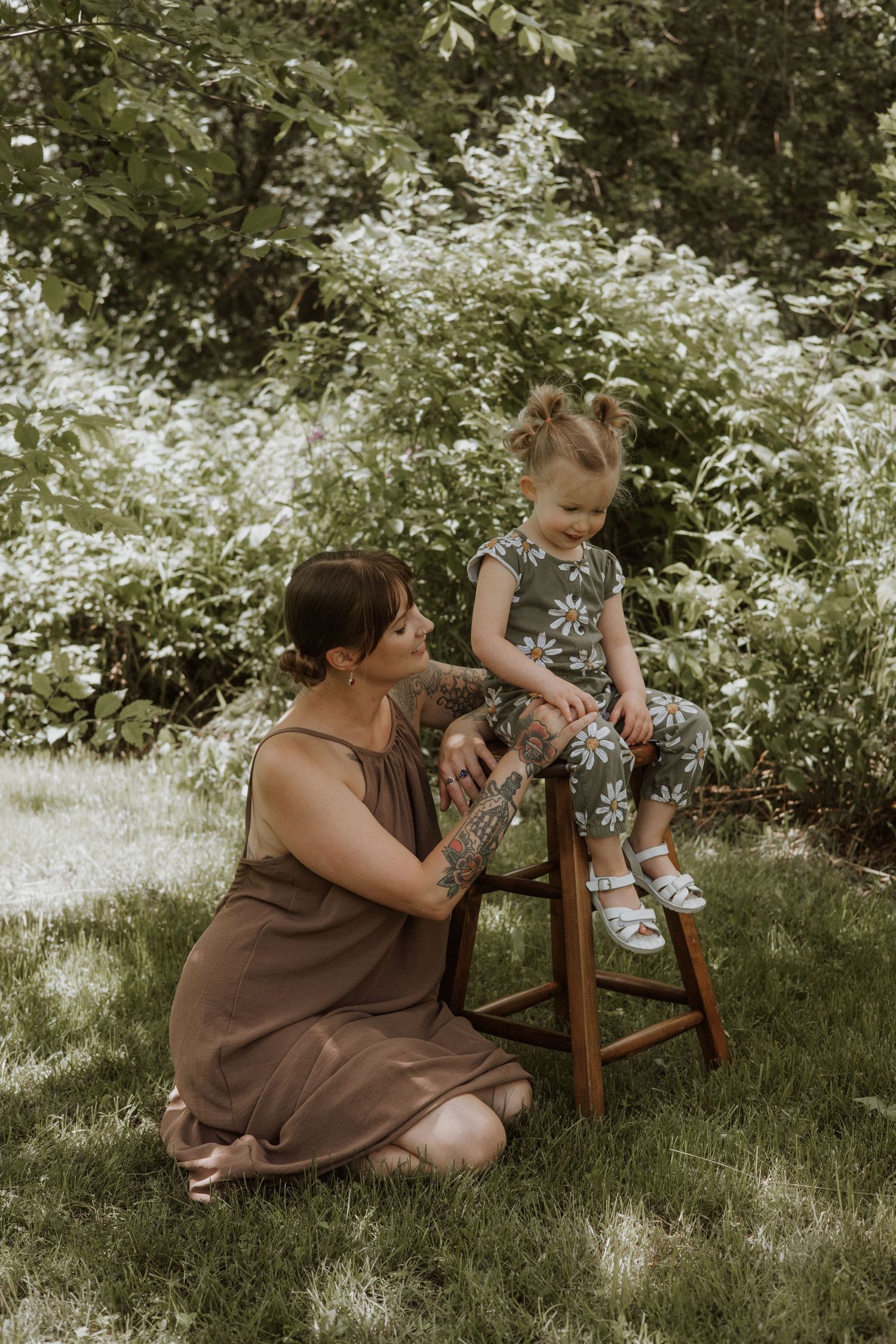 A tattooed mother kneels on grass interacting with her toddler daughter sitting on a wooden stool outdoors.