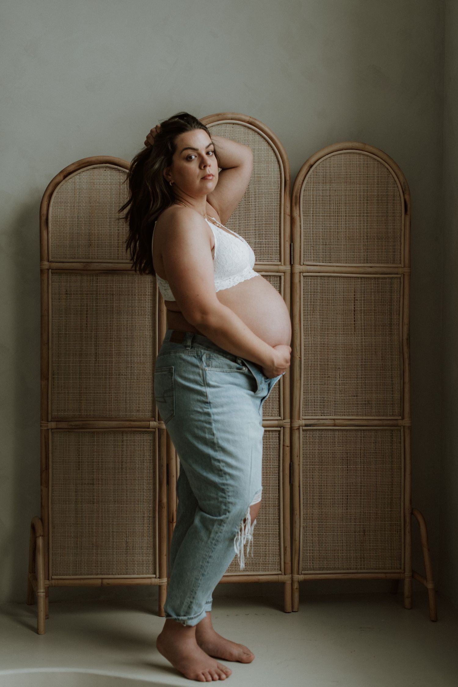 Pregnant woman in white lace bra and distressed jeans standing before a rattan folding screen.