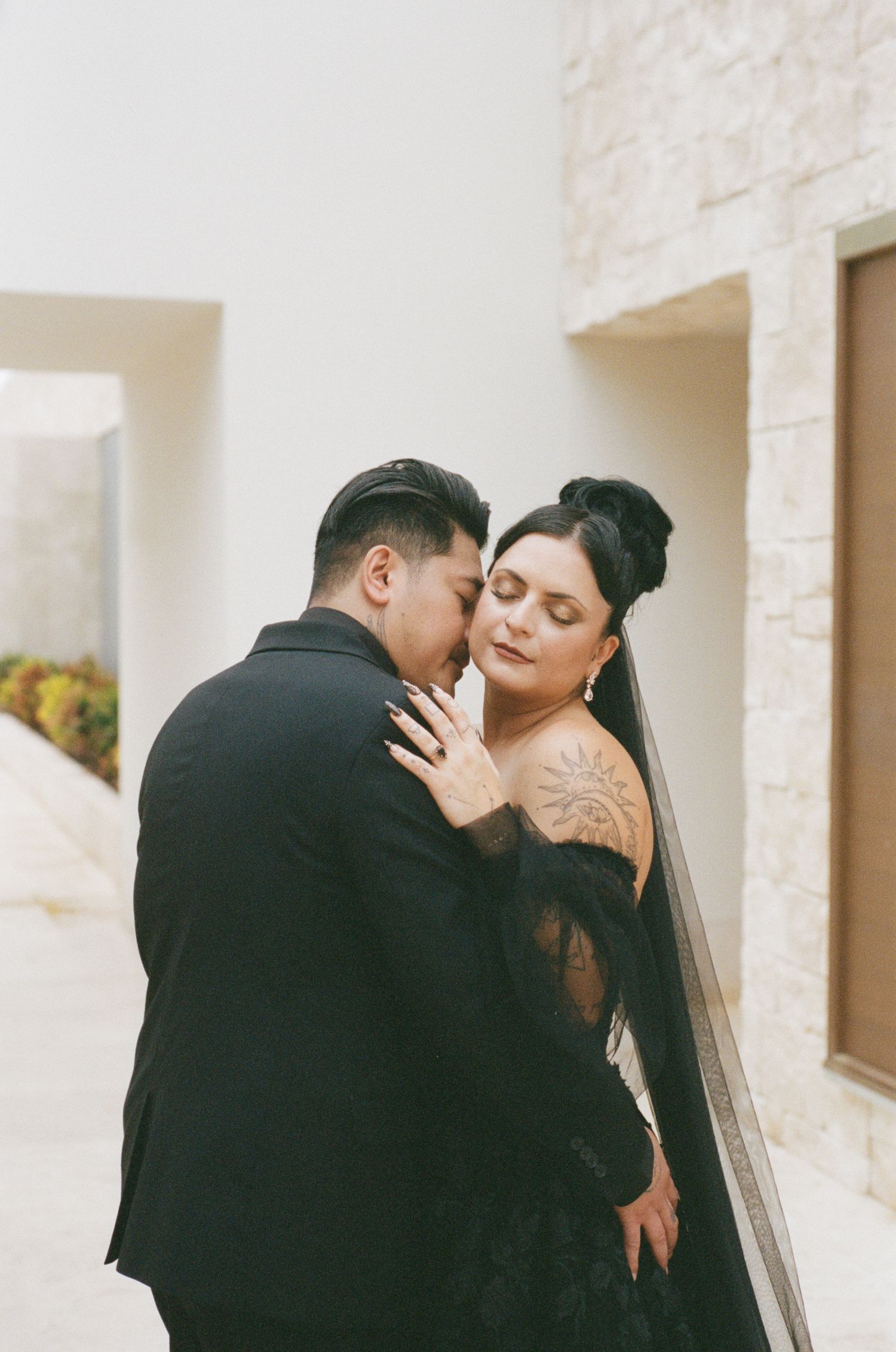 Bride with tattoo in black wedding dress embraces groom in black suit at modern outdoor venue.