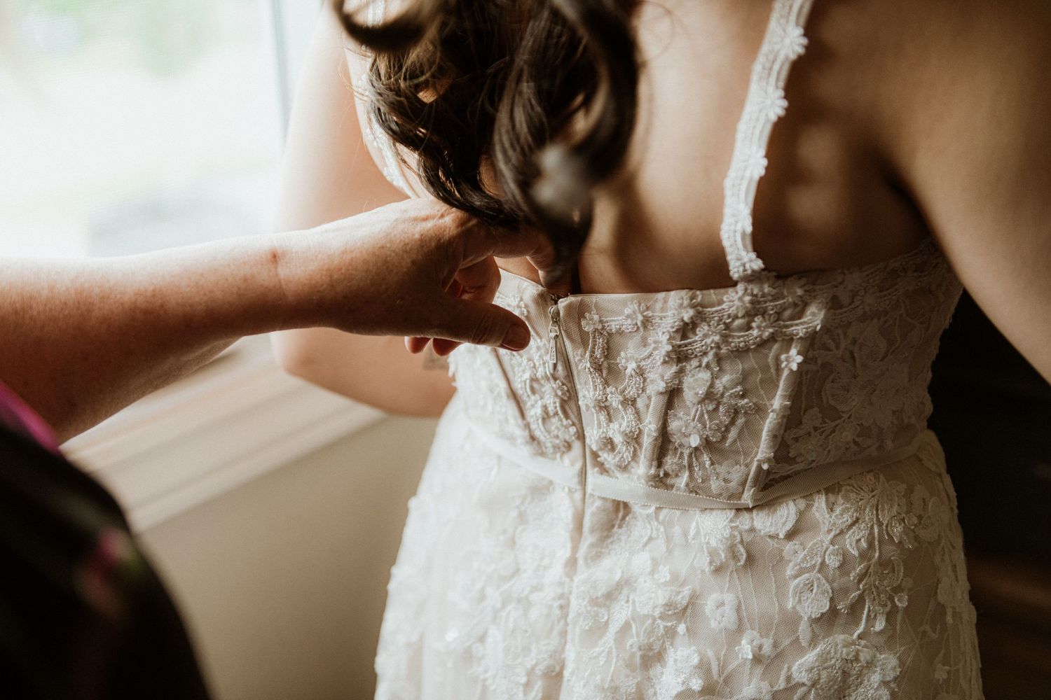 Close-up of bridesmaid fastening delicate buttons on back of bride's lace wedding dress.