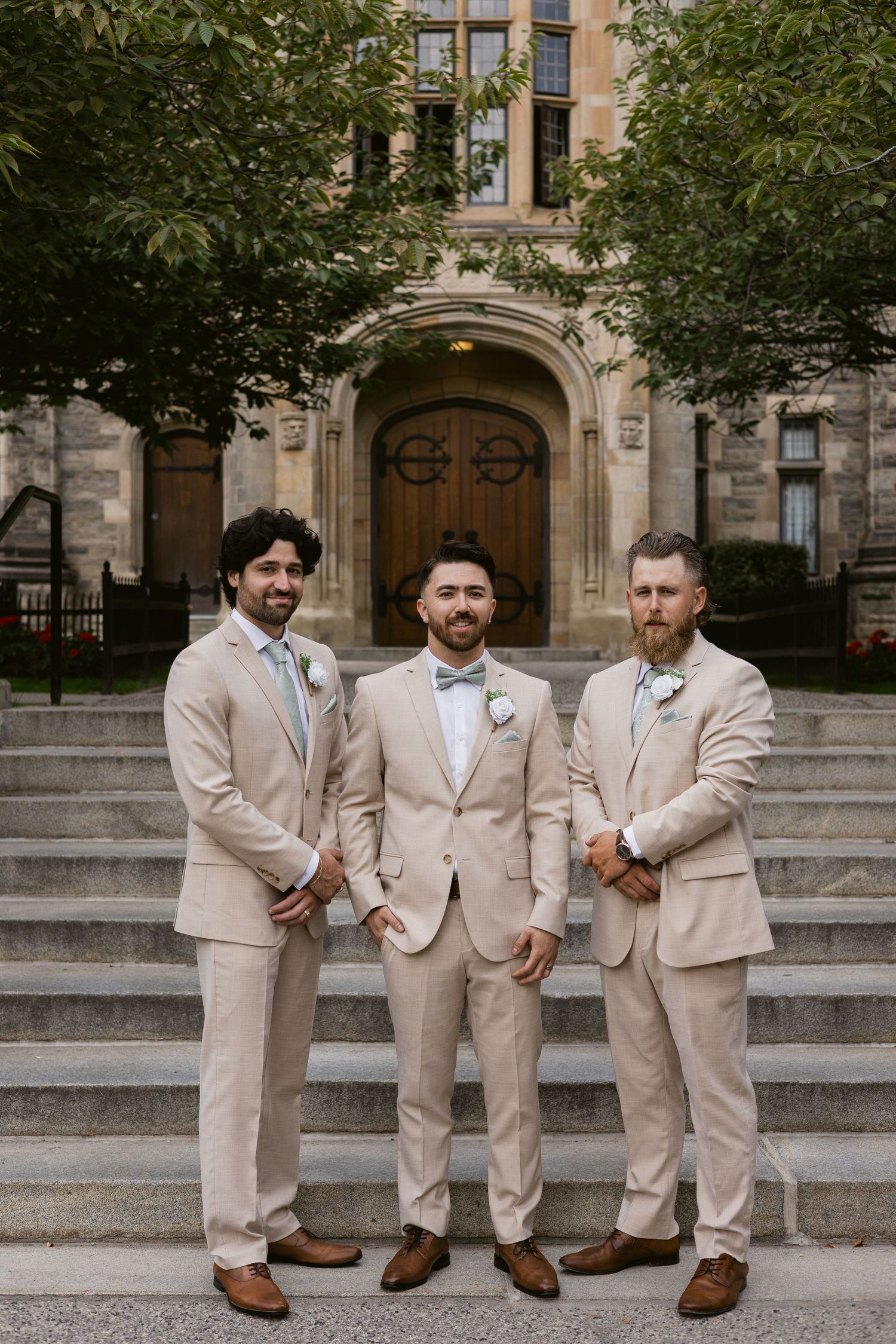 Three groomsmen in matching tan suits with bow ties stand on stone steps outside historic building.