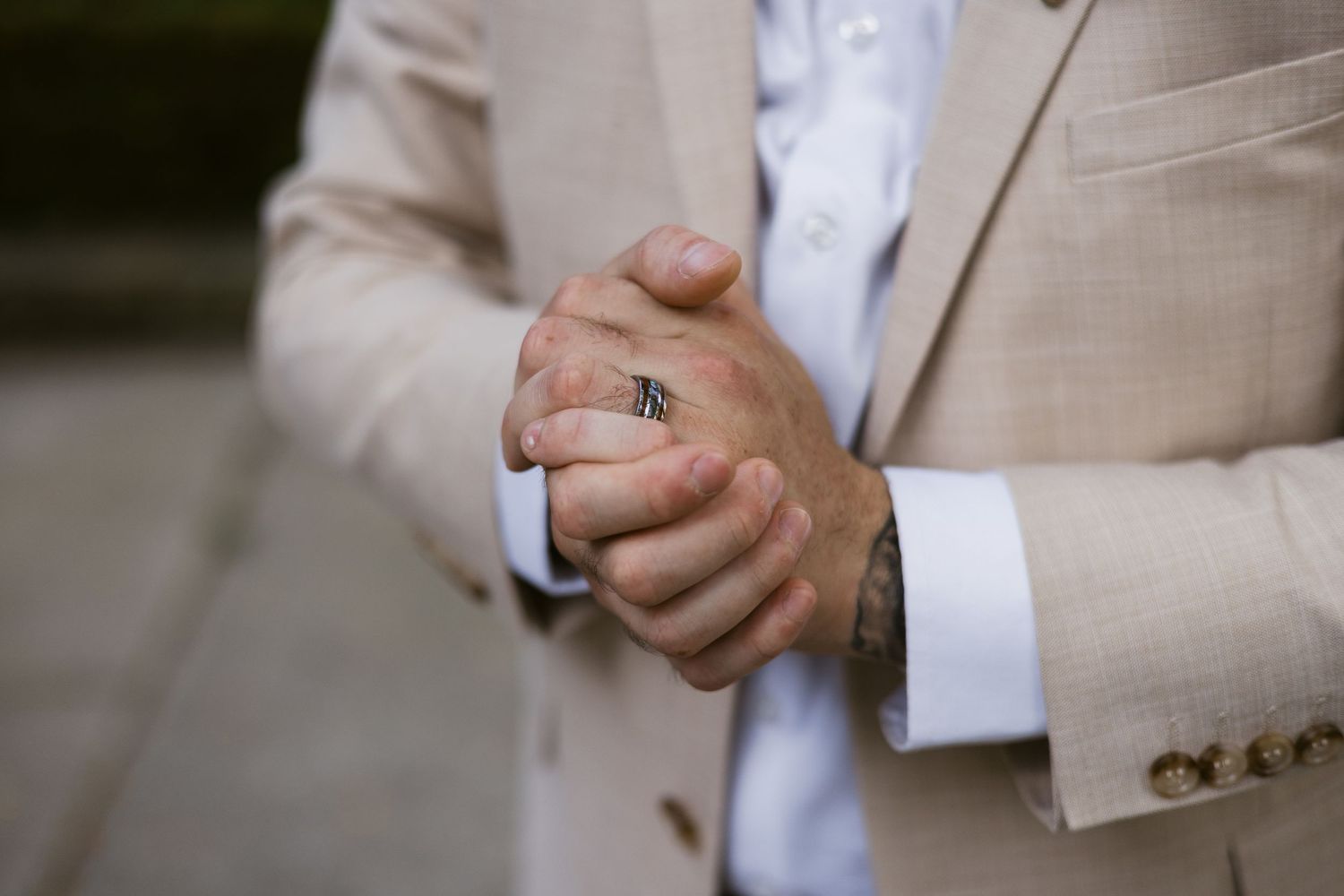 Close-up of groom's hands clasped together wearing a ring, beige suit with white dress shirt.