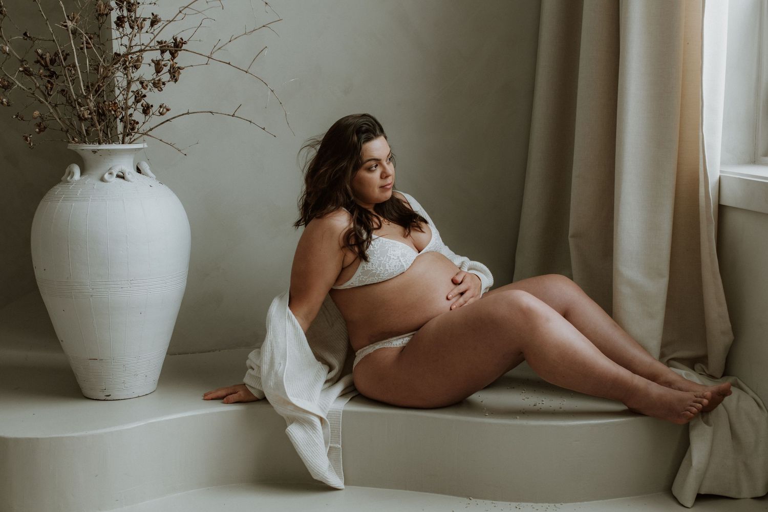 Pregnant woman in white lingerie sitting on a raised ledge next to a large white decorative vase.