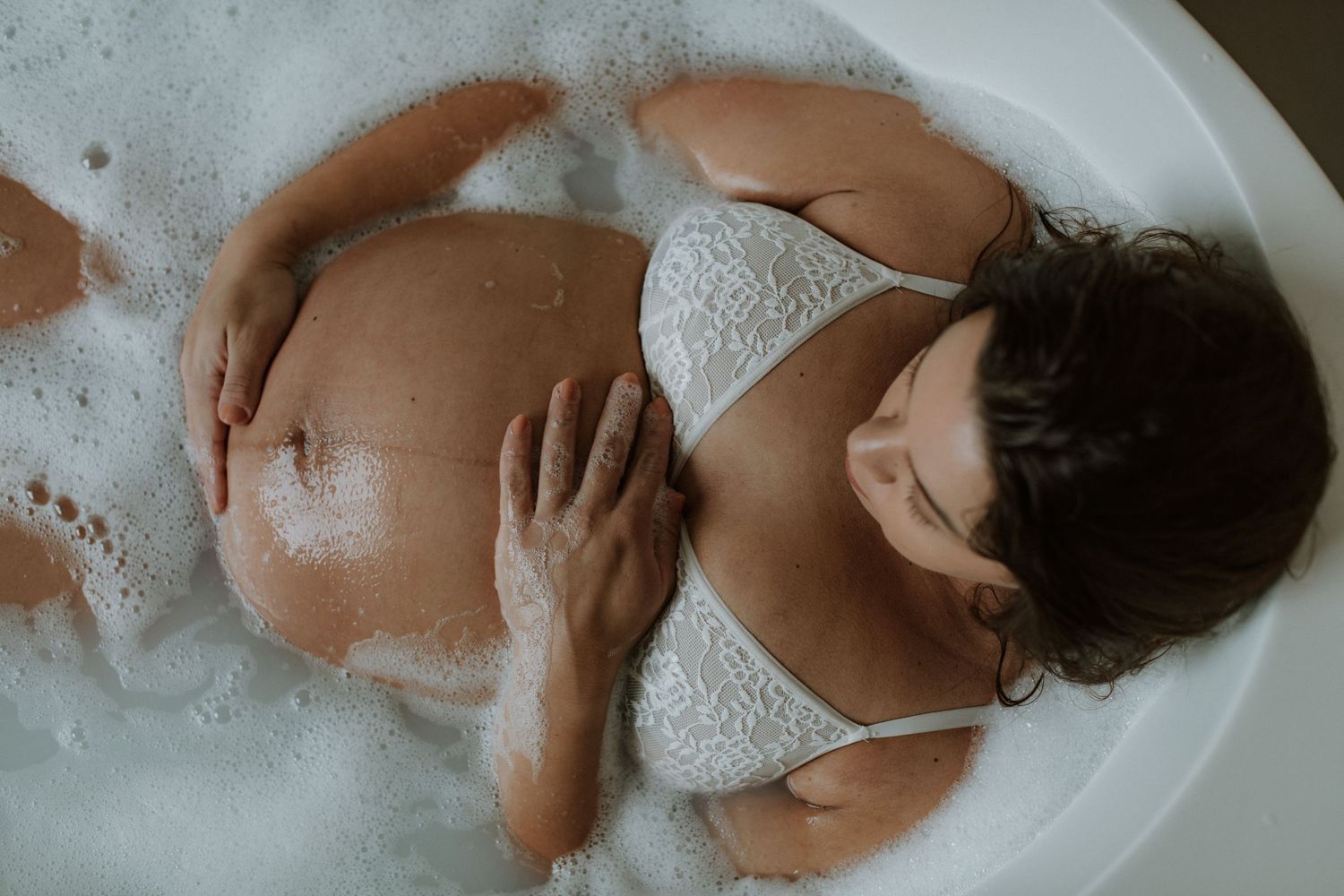 Overhead view of a pregnant woman in white lace lingerie soaking in a bubble bath.