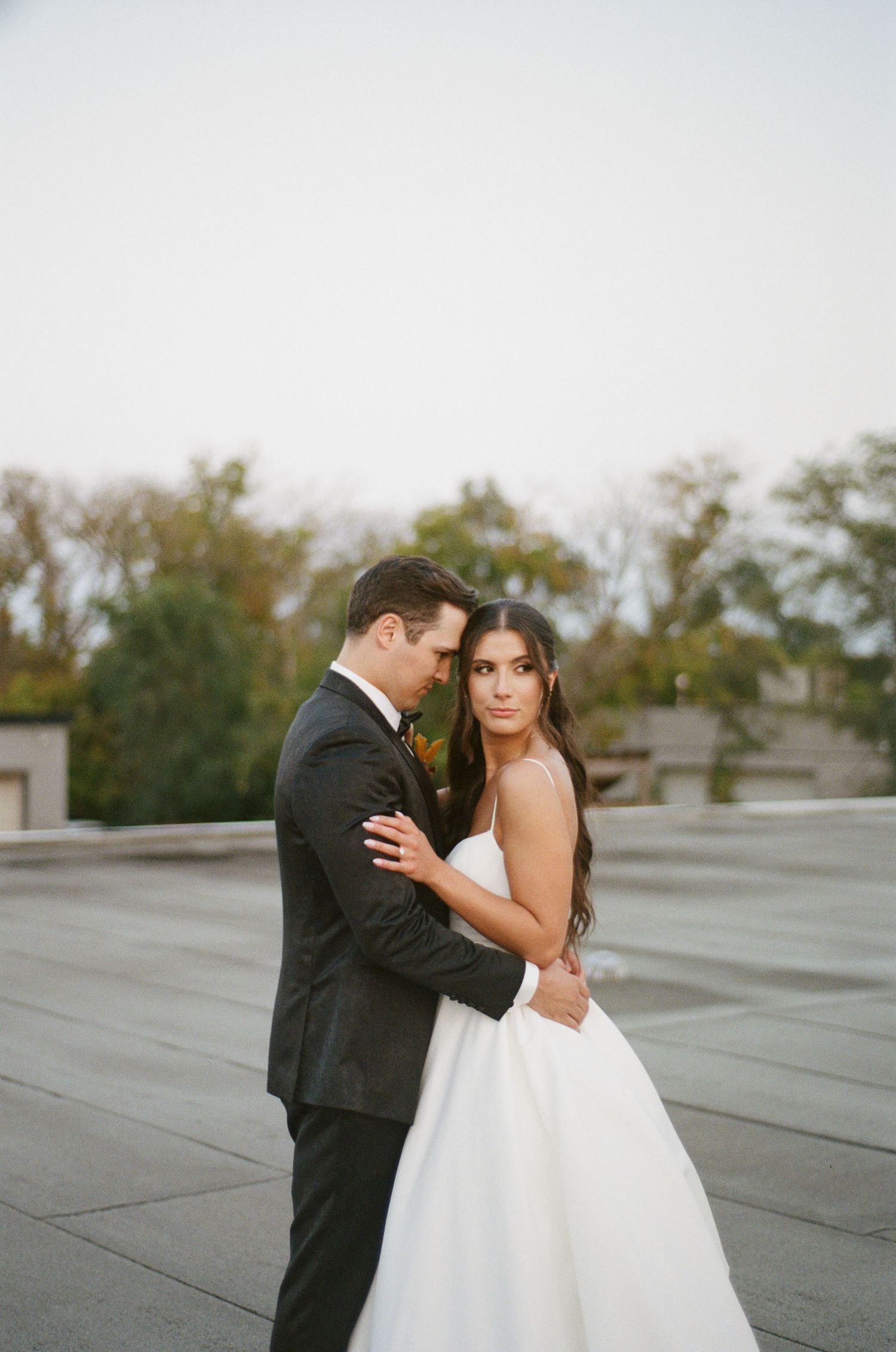 Bride in spaghetti strap gown gazes at camera while groom holds her close on rooftop.