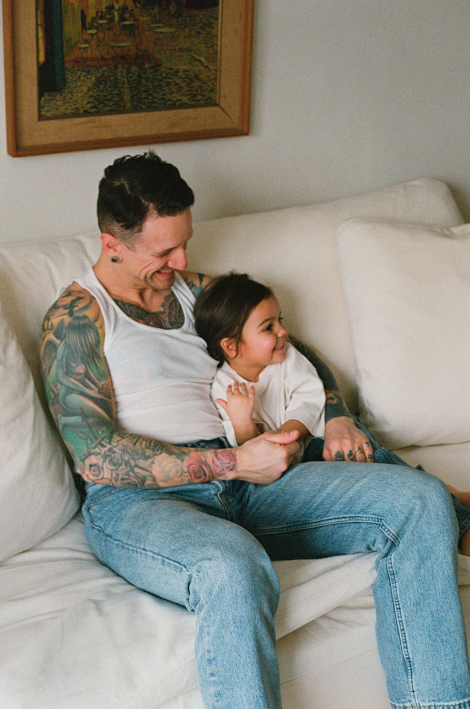 Heavily tattooed father in white tank top smiling and bonding with baby on white sofa.
