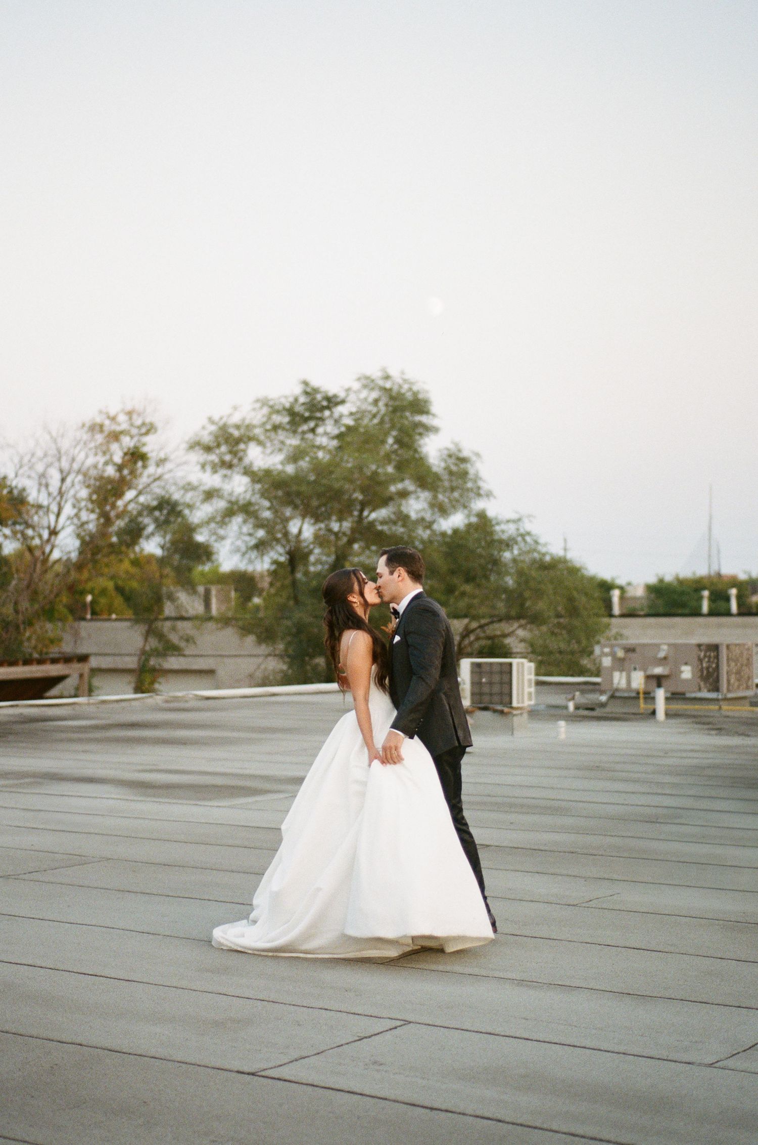Bride and groom share a tender kiss on rooftop venue with moon visible in soft evening sky.