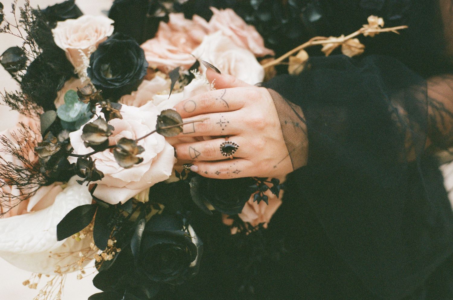 Bride\u2019s tattooed hands with rings touching dark and white roses in wedding bouquet.