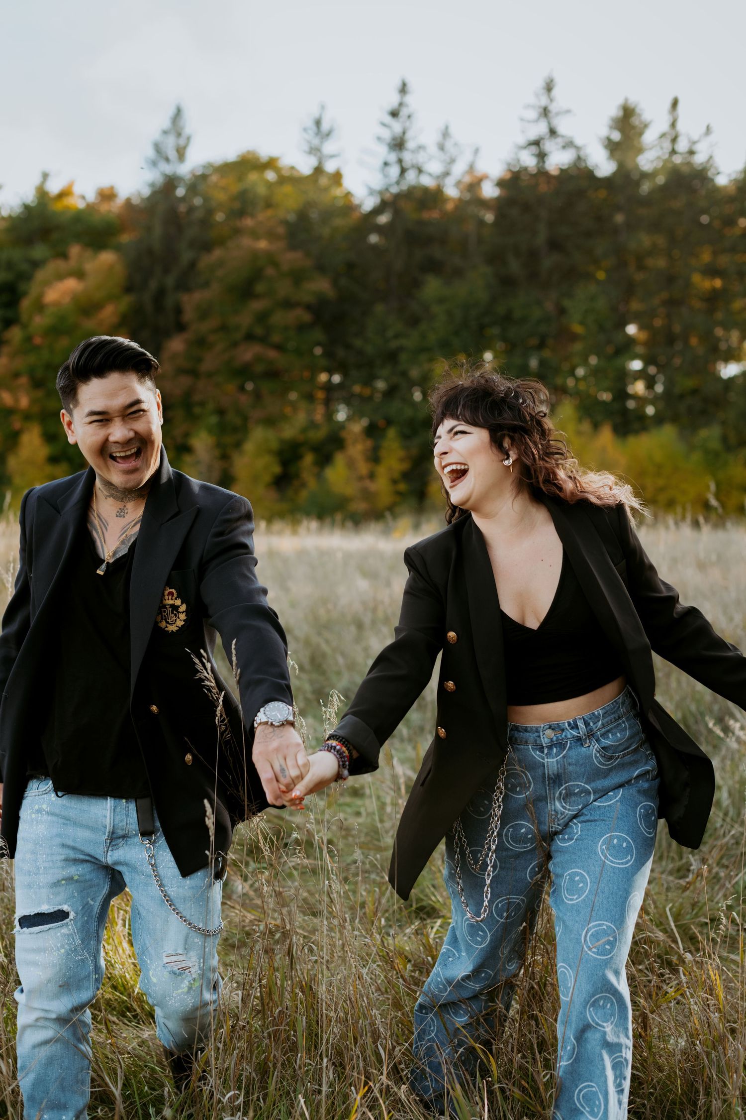 Joyful couple laughing together while holding hands and walking through autumn field with colorful trees in background.