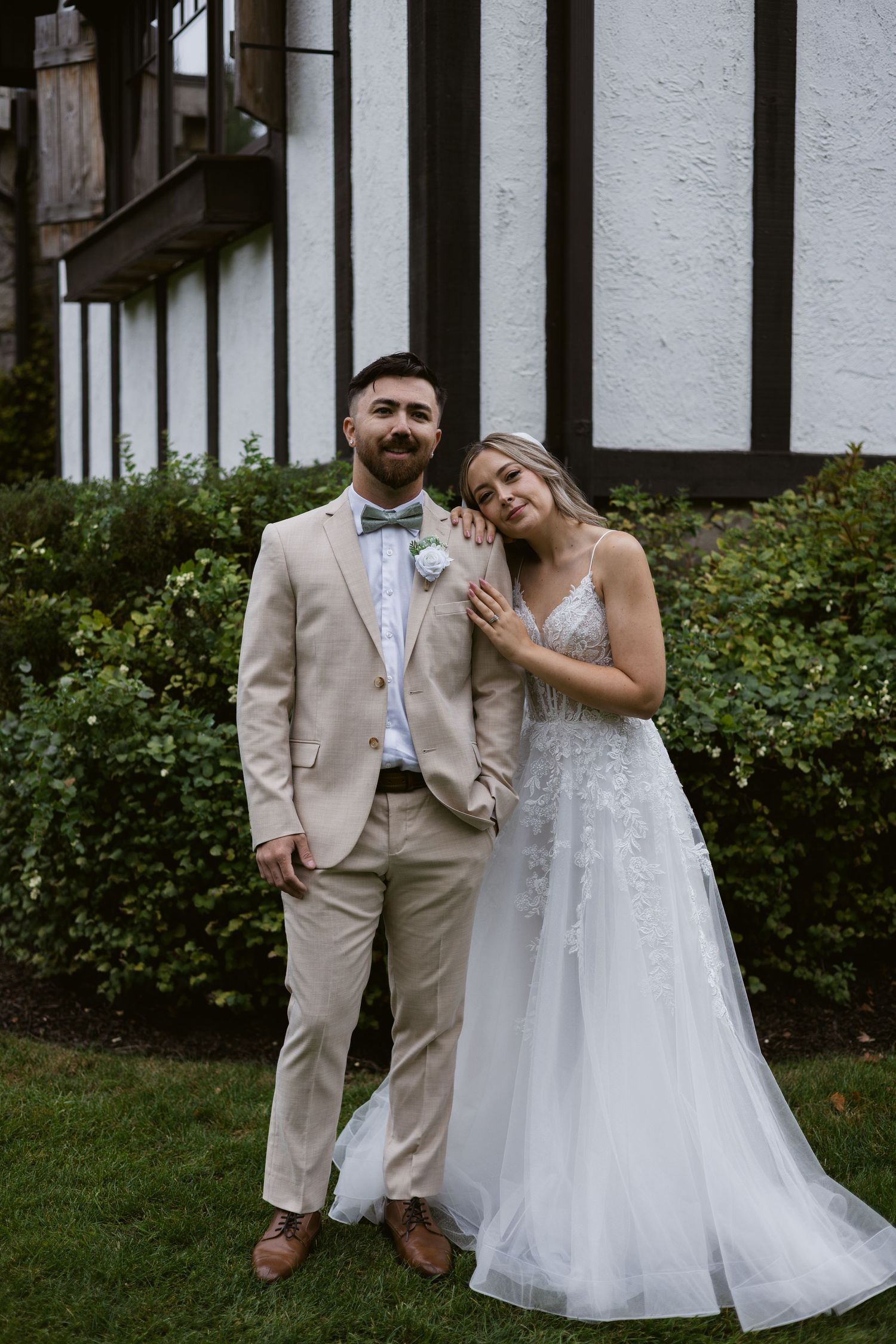 Bride leaning on grooms shoulder posing in front of Tudor-style building on wedding day.