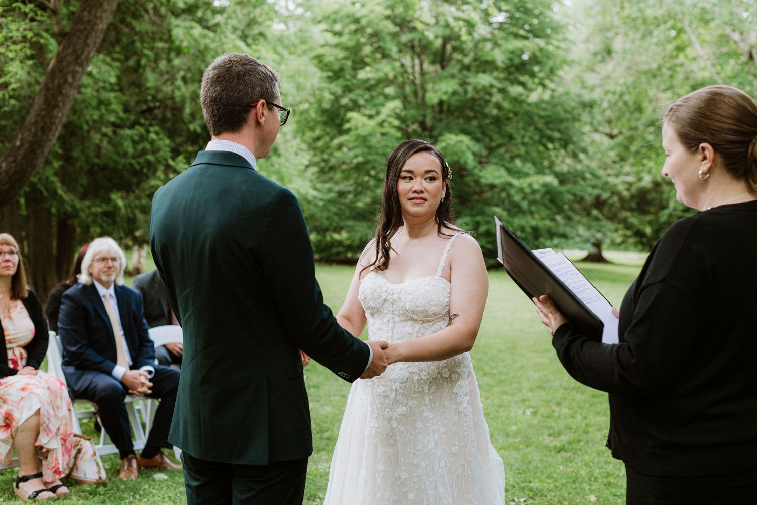 Bride in lace gown holds grooms hands smiling during outdoor wedding ceremony with officiant nearby.