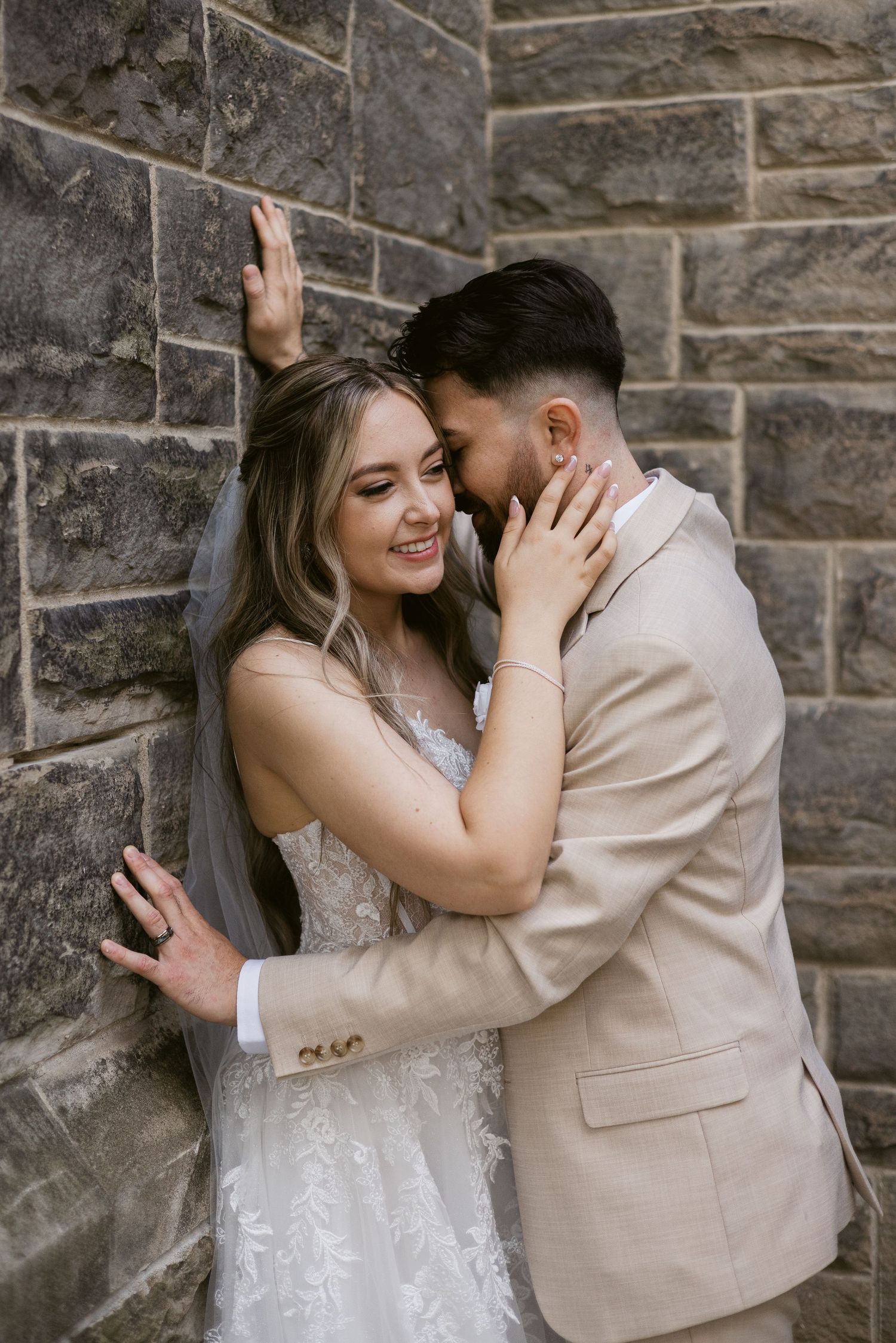 Laughing bride leans against stone wall as groom in tan suit nuzzles her during wedding portraits.