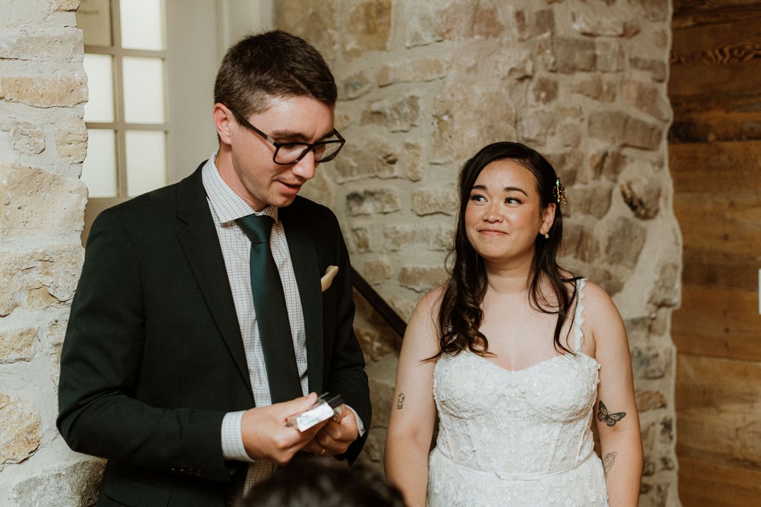 Groom reads vows while bride listens during intimate wedding ceremony near stone wall.
