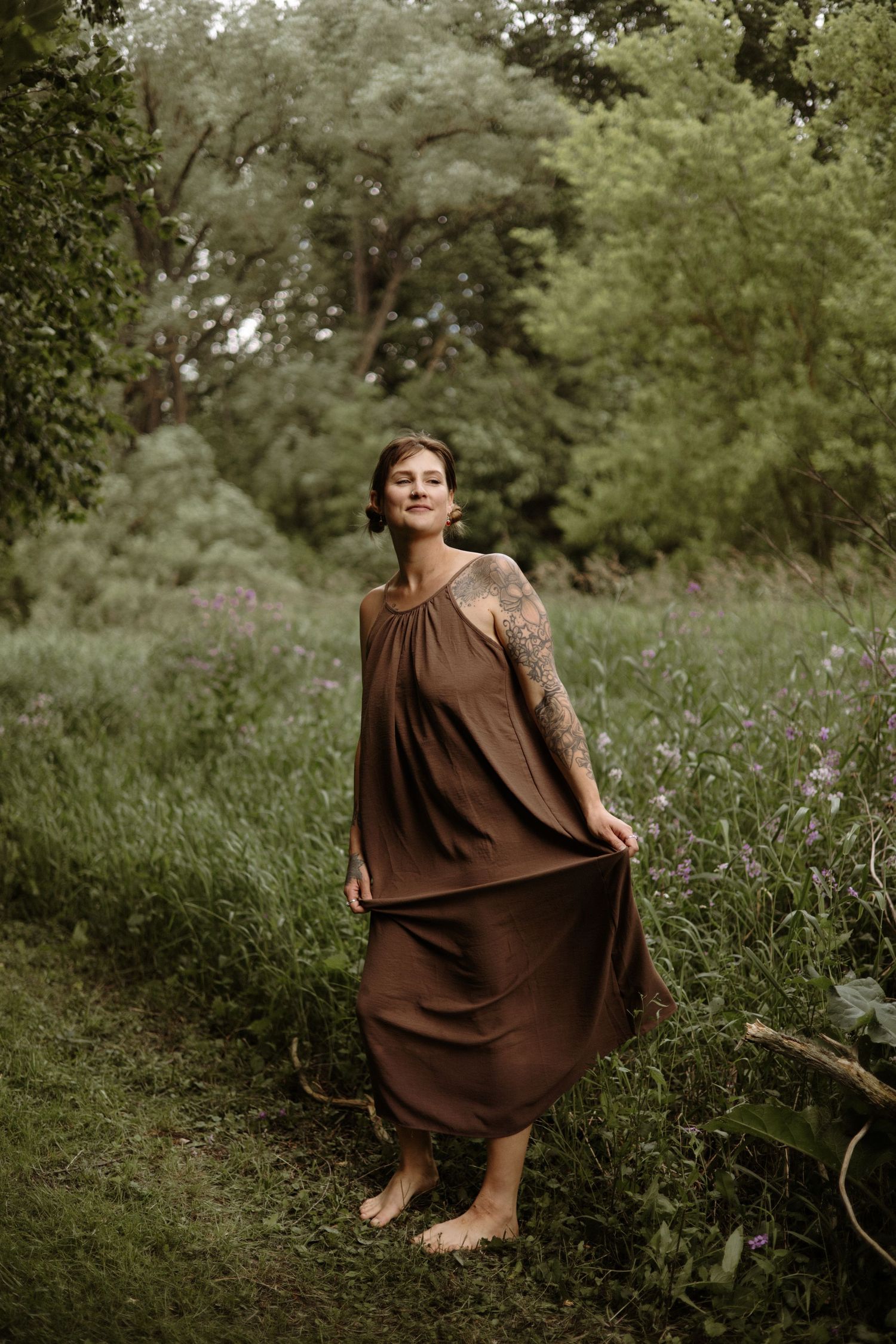 A smiling barefoot tattooed woman in a flowing brown dress stands among wildflowers in a lush green forest.